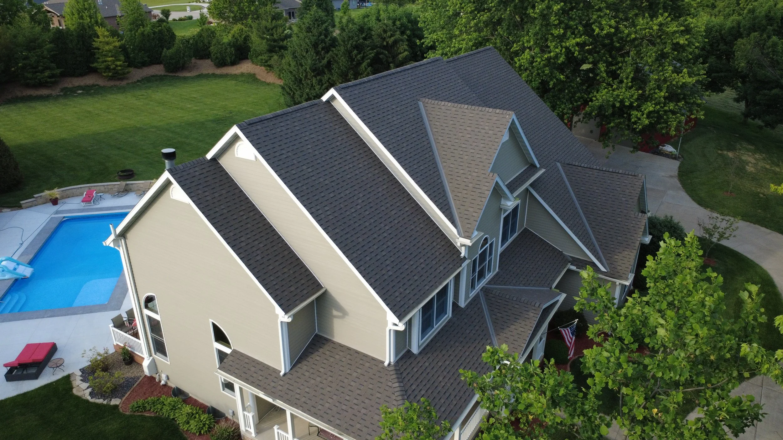 An aerial view of a multi-story house with a complex roof design, a backyard with a swimming pool, outdoor furniture, and surrounding green trees and lawn.