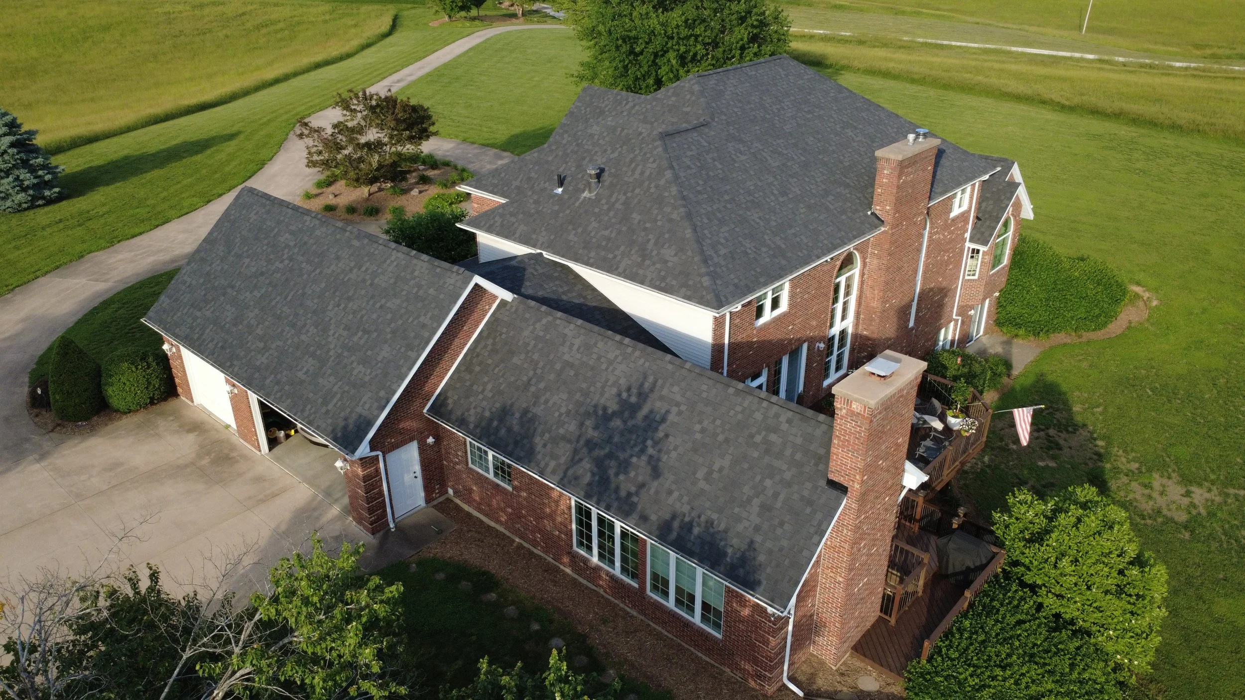 An aerial view of a large brick house with a dark gray shingled roof, surrounded by green lawns, trees, and a curved driveway.