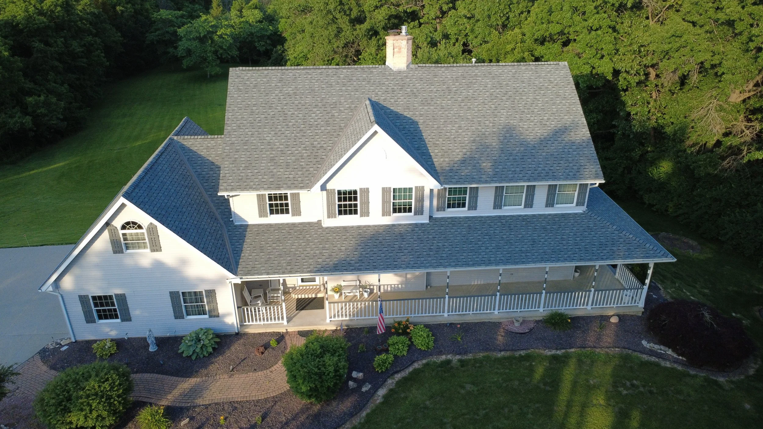 Aerial view of a large white house with a gray shingled roof, surrounded by green trees, a lawn, and a landscaped garden with bushes and flowering plants.