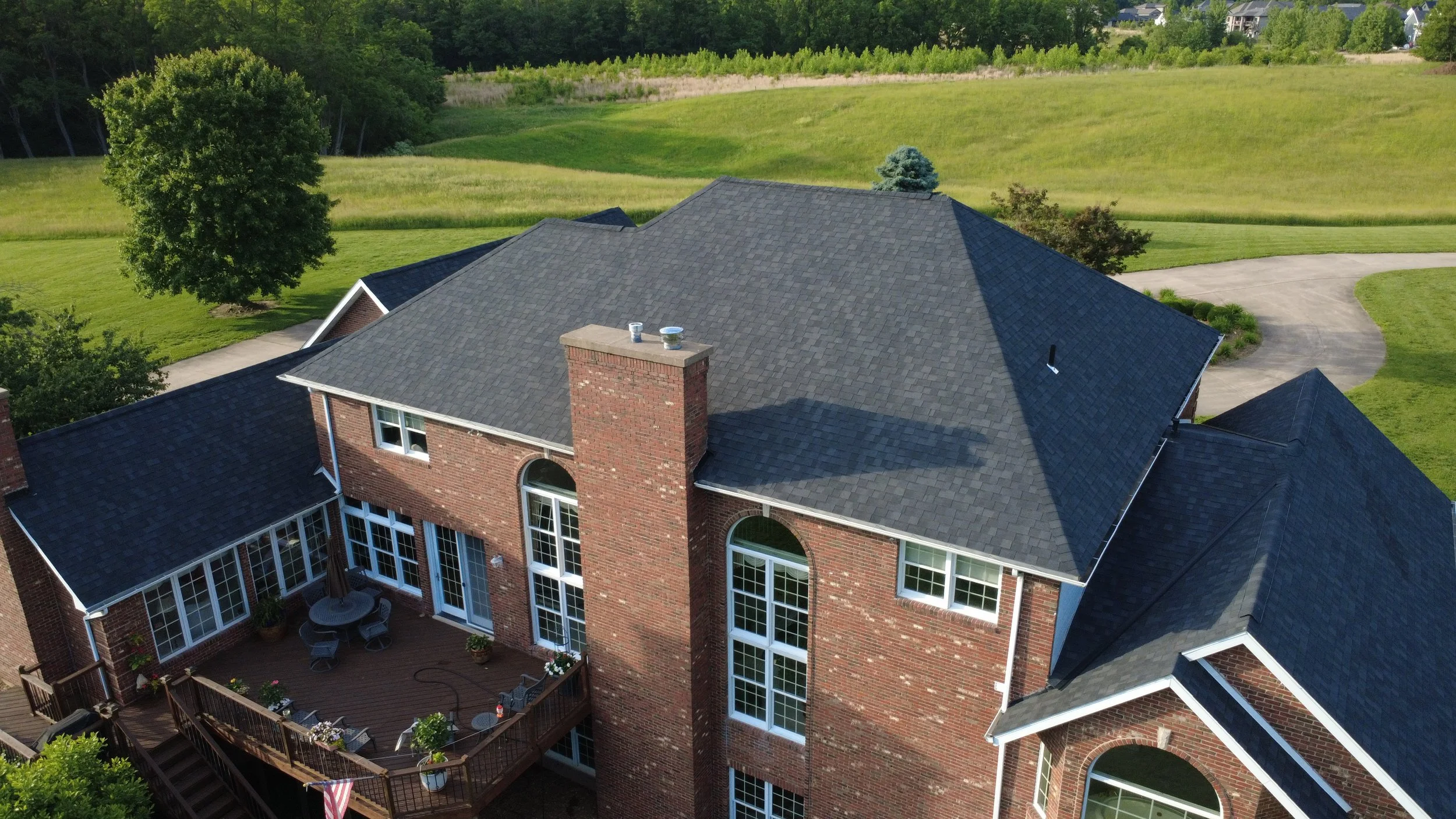 Aerial view of a large brick house with a dark gray shingle roof, overlooking a lush green backyard with a deck, patio furniture, and surrounding trees and open fields.