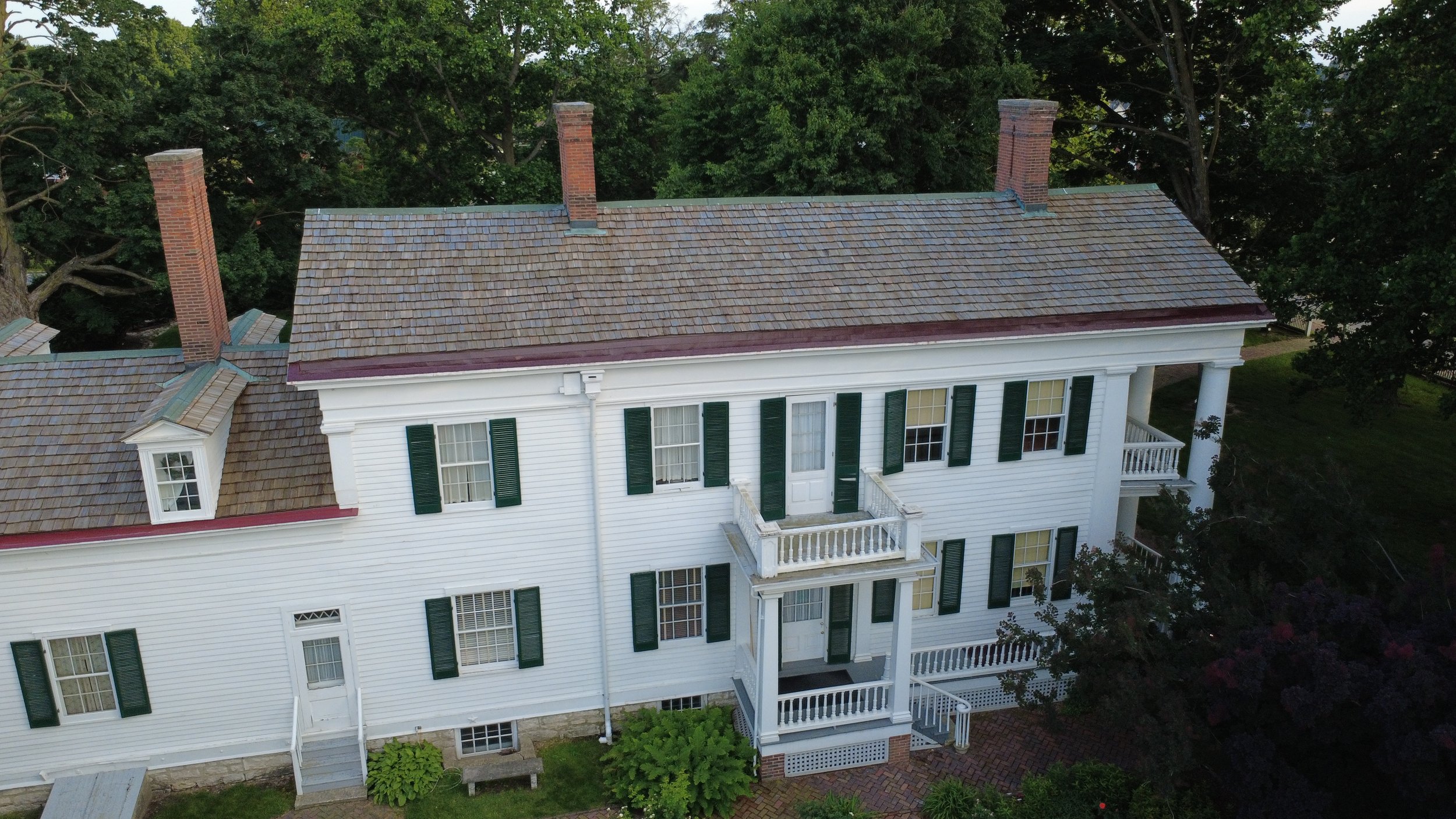 Aerial view of a large white house with multiple chimneys and green shutters, surrounded by trees.
