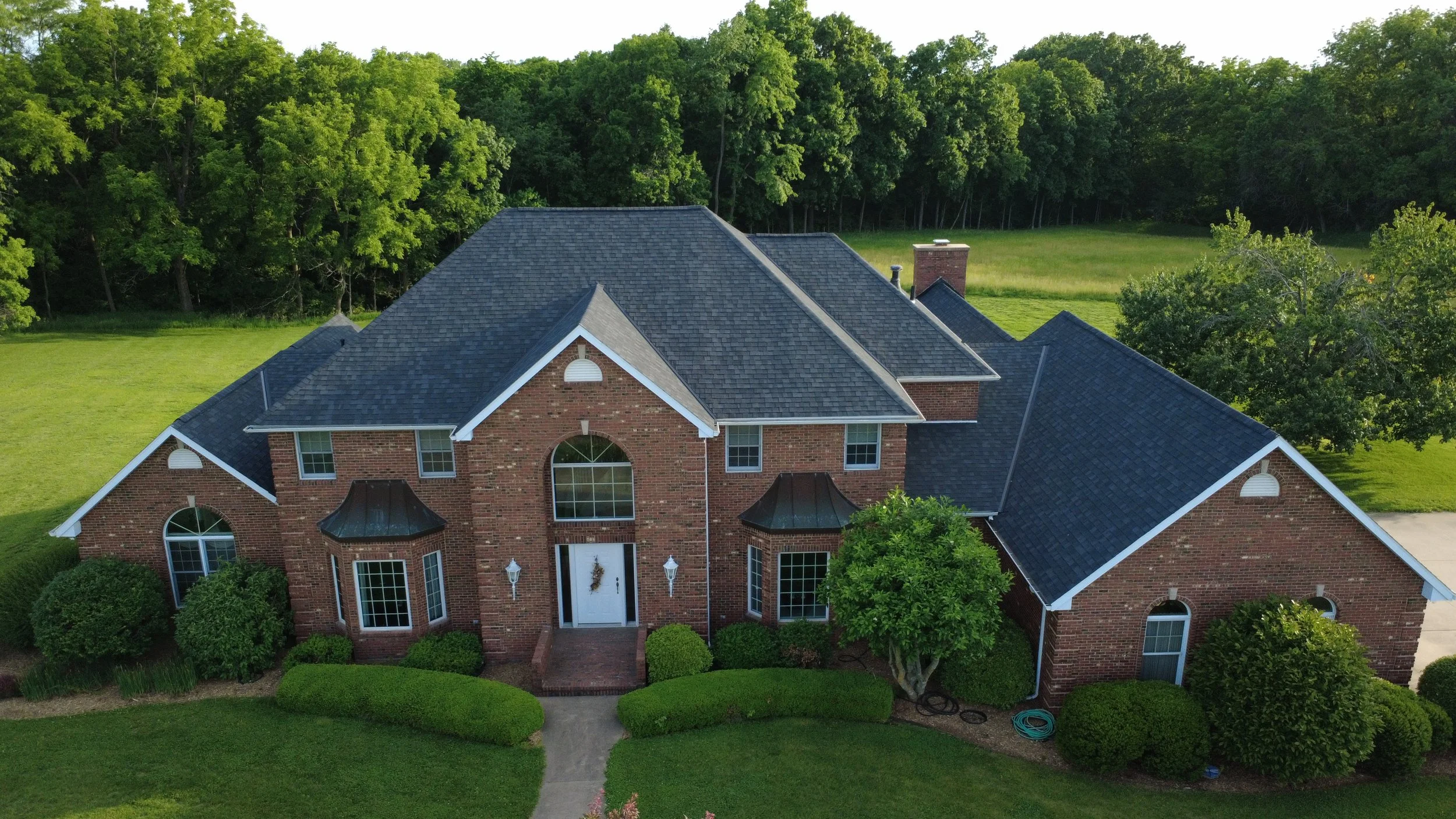 Aerial view of a large brick house with multiple rooflines, arched windows, and a front door with a small porch, surrounded by green trees and a well-manicured lawn.
