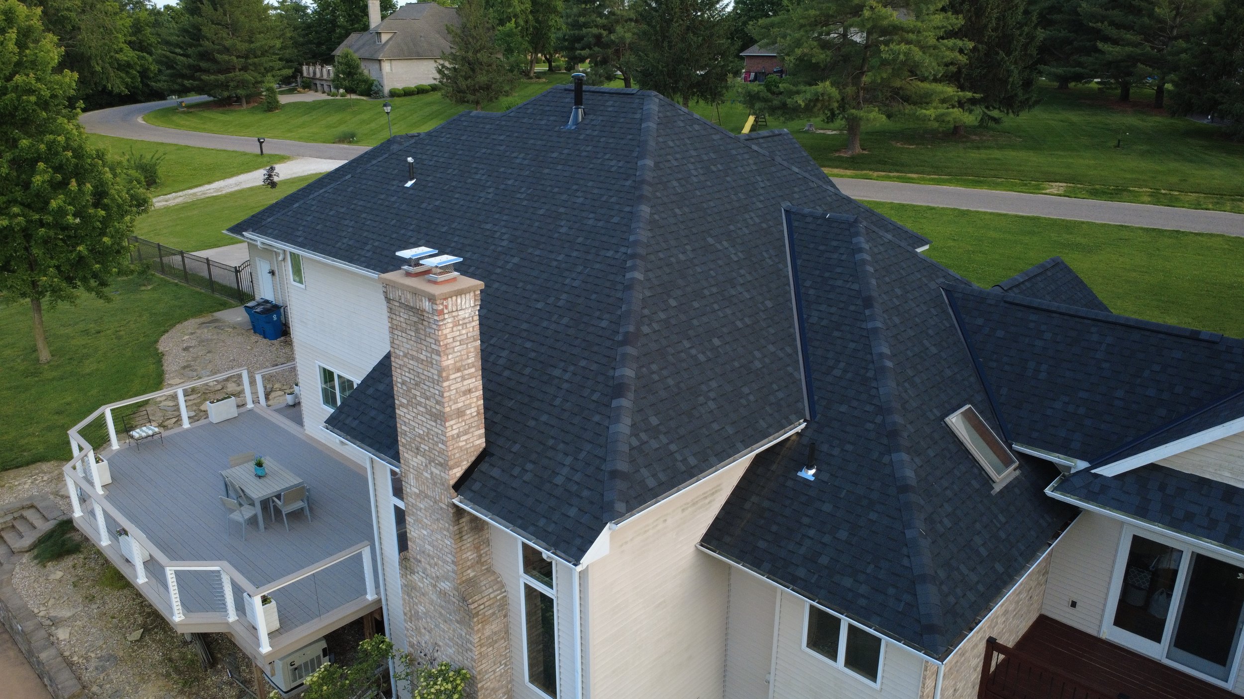 Aerial view of a house with a dark gray shingled roof, brick chimney, and white exterior walls, surrounded by green yard and trees.