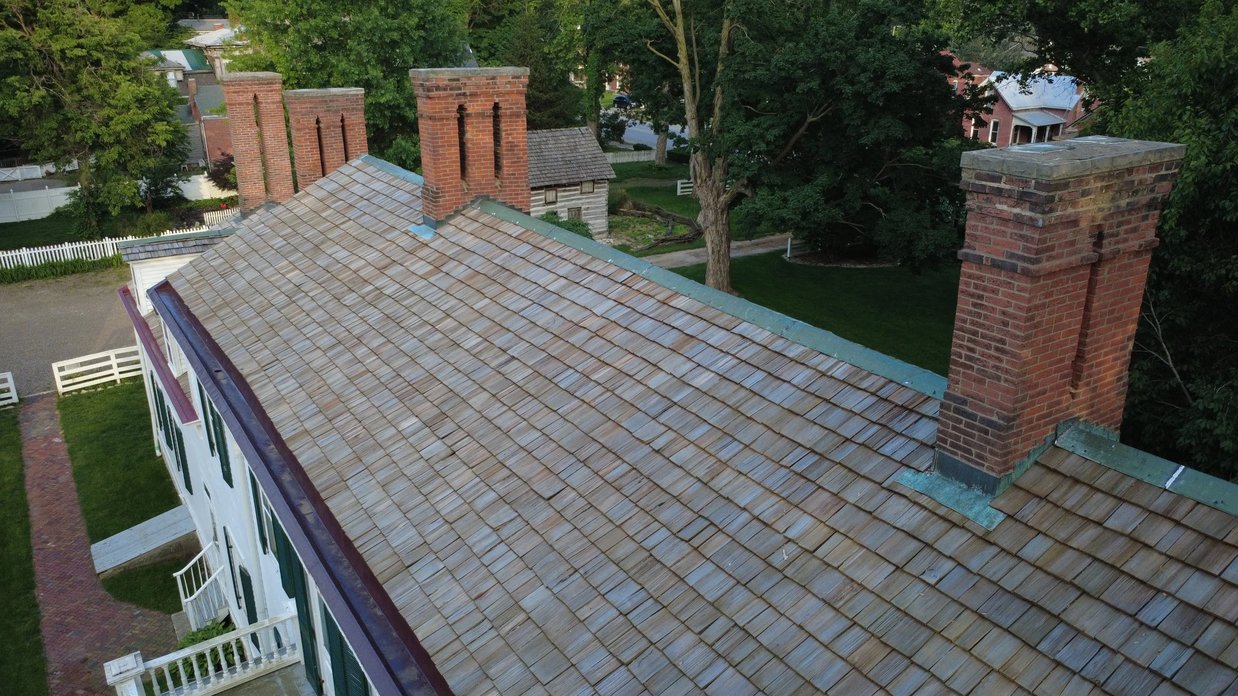 An aerial view of a house with a tiled roof and multiple brick chimneys, surrounded by lush green trees and residential neighborhoods.