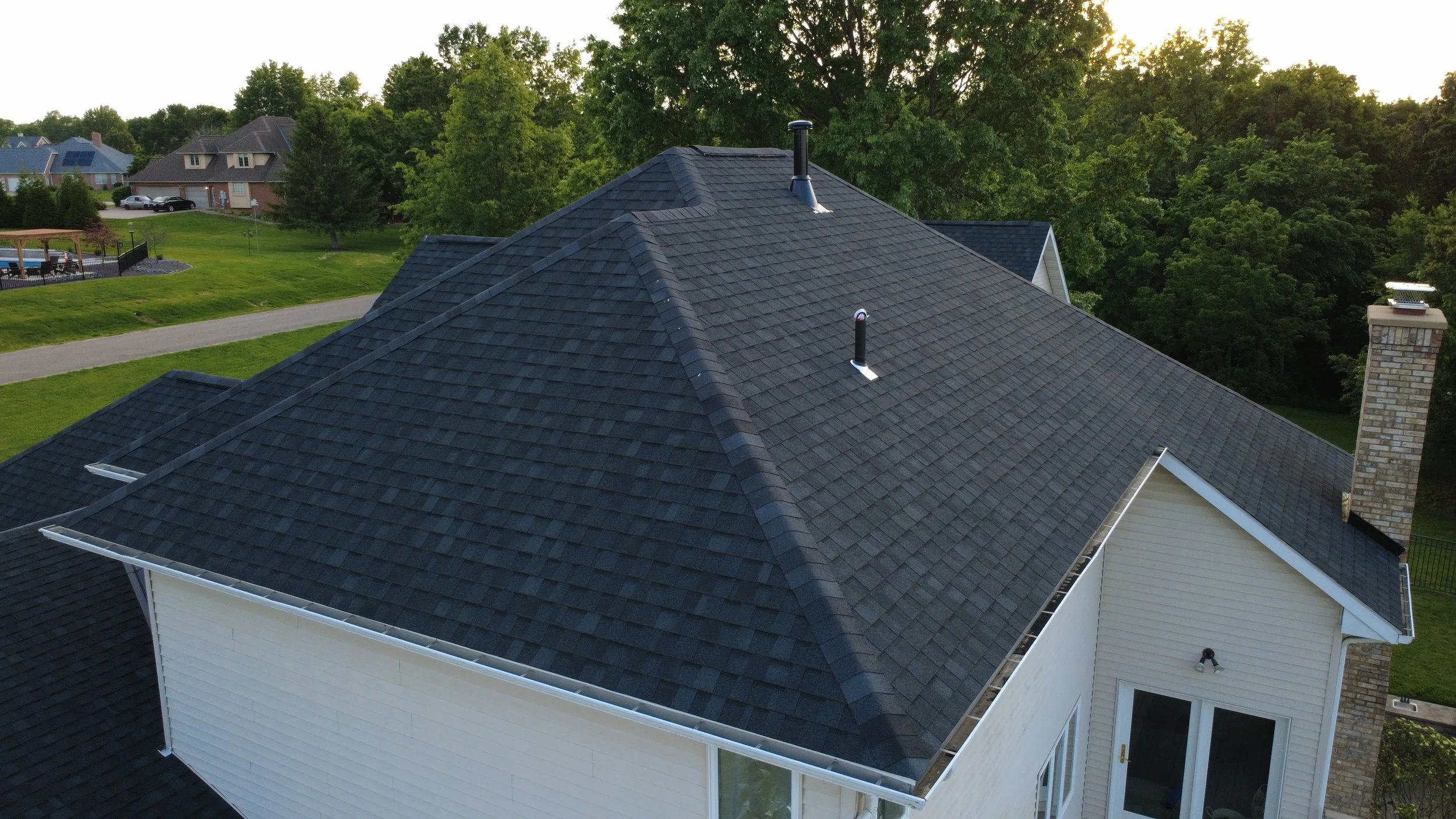 Aerial view of a house roof with dark shingles, two vent pipes, and a chimney, surrounded by trees and neighboring houses.