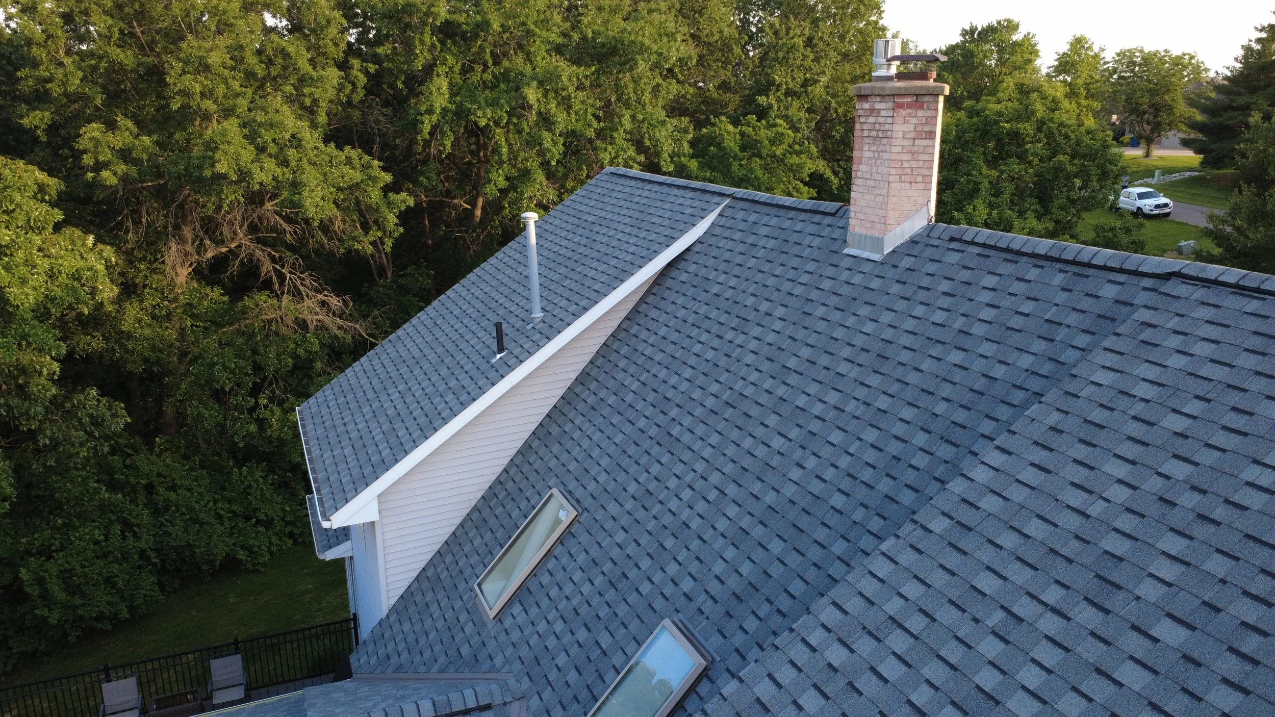 A house roof with gray shingles, a chimney, and skylights, surrounded by green trees and a neighboring yard with a parked white vehicle.