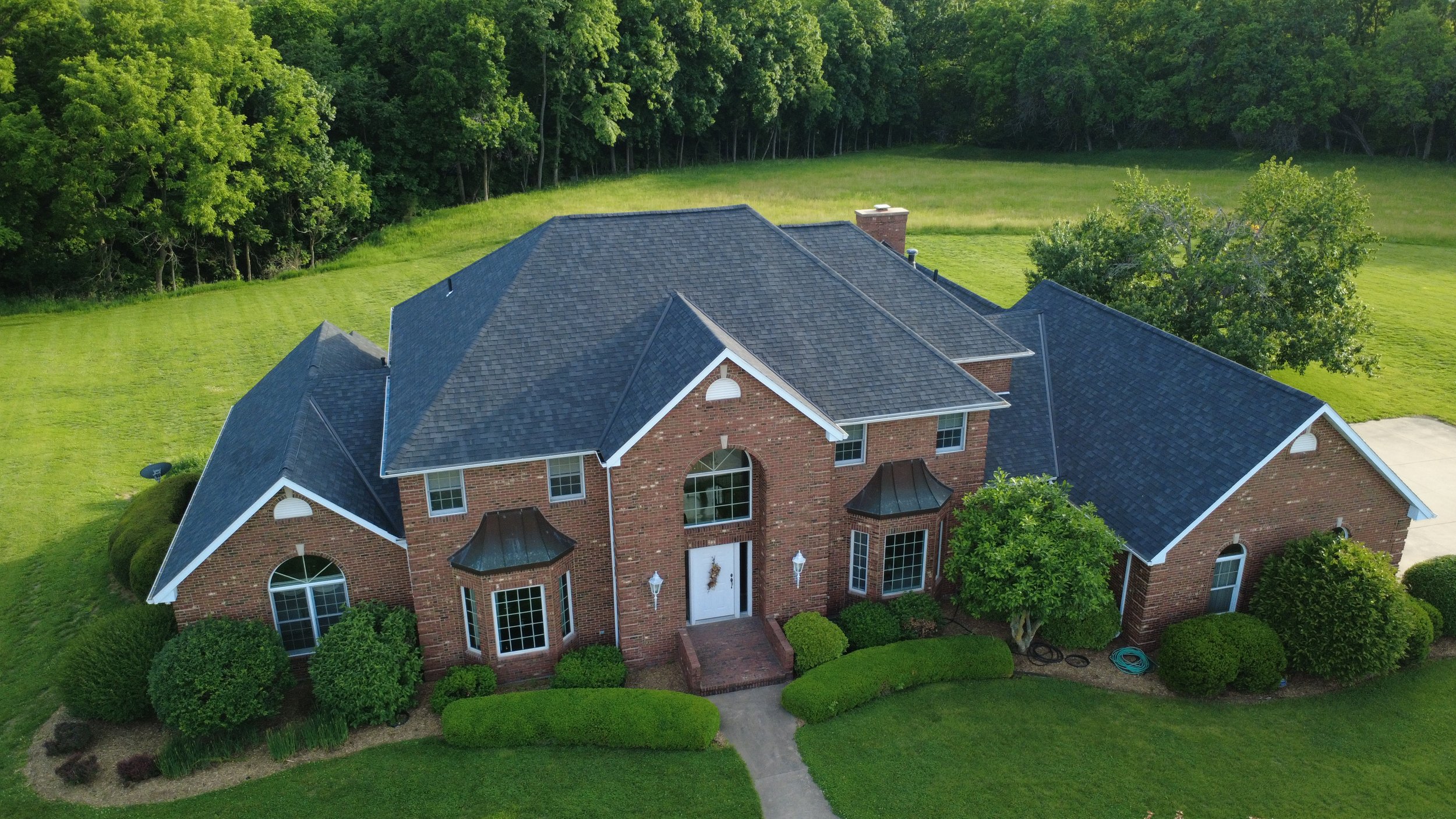 A large brick house with multiple gabled roofs, surrounded by a lush green yard and trees, with a concrete driveway on the right.