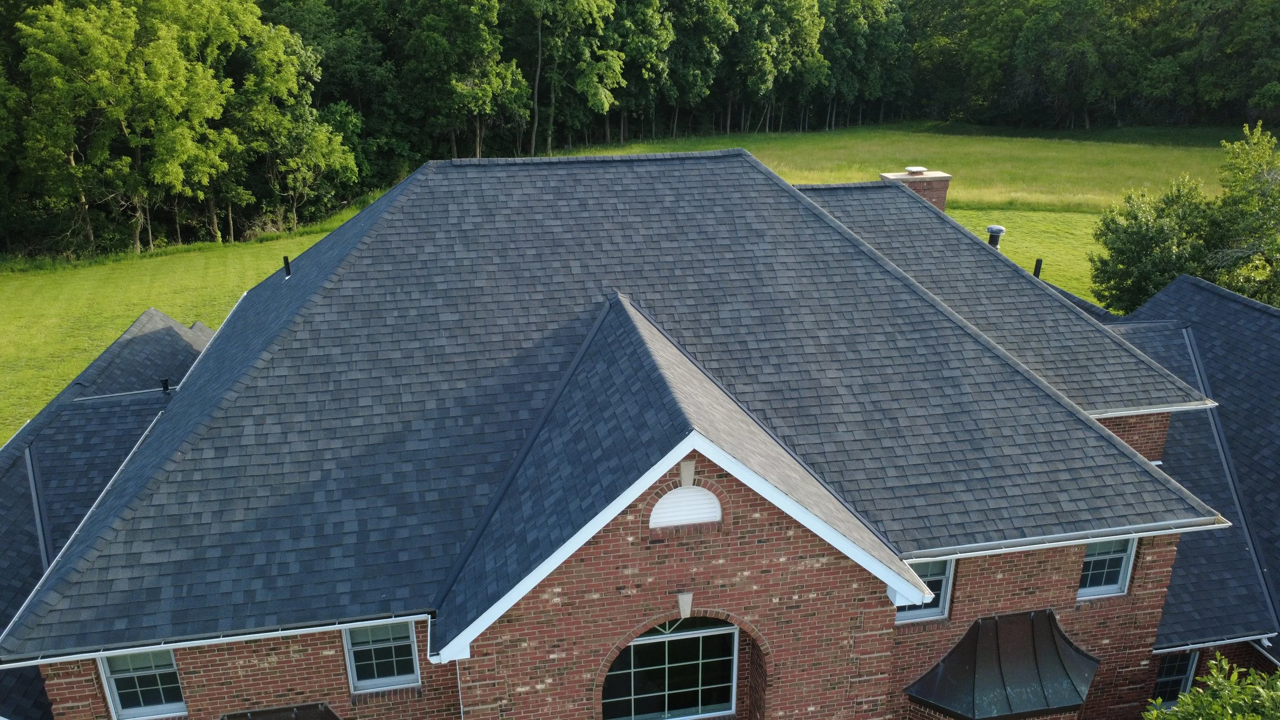 Aerial view of a house with a dark gray shingle roof, brick walls, multiple chimneys, and surrounding green yard and trees.