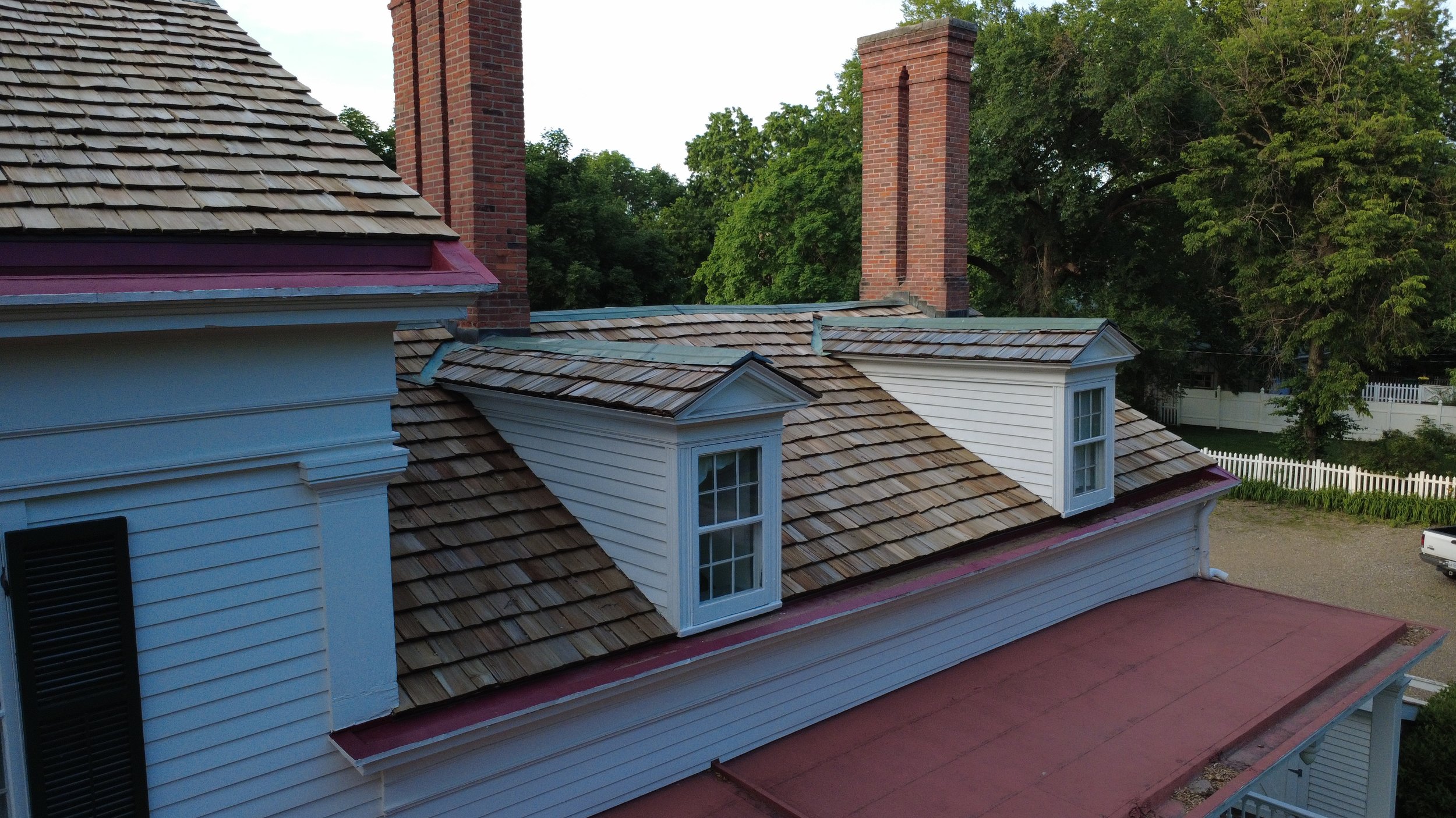 View of a house's roof with shingled and tiled sections, two prominent brick chimneys, and trees in the background.