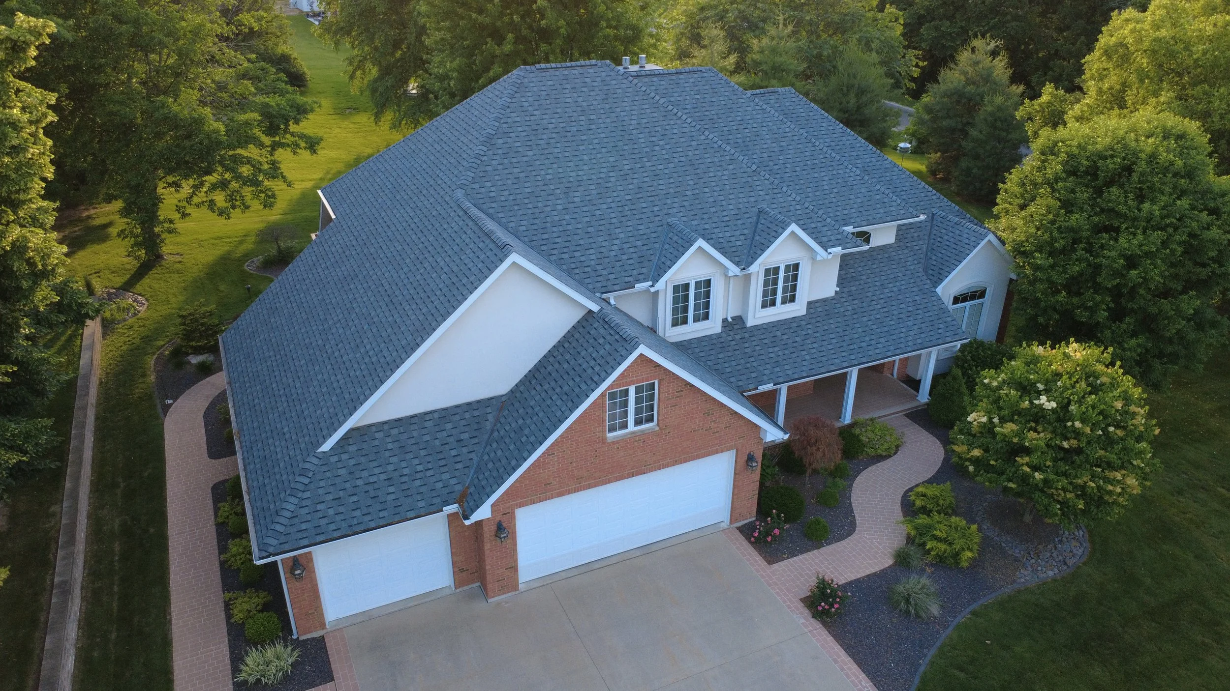 An aerial view of a two-story house with a large gray shingle roof, brick and white exterior walls, multiple windows, a garage, and a landscaped yard with trees, shrubs, and a curved brick pathway.