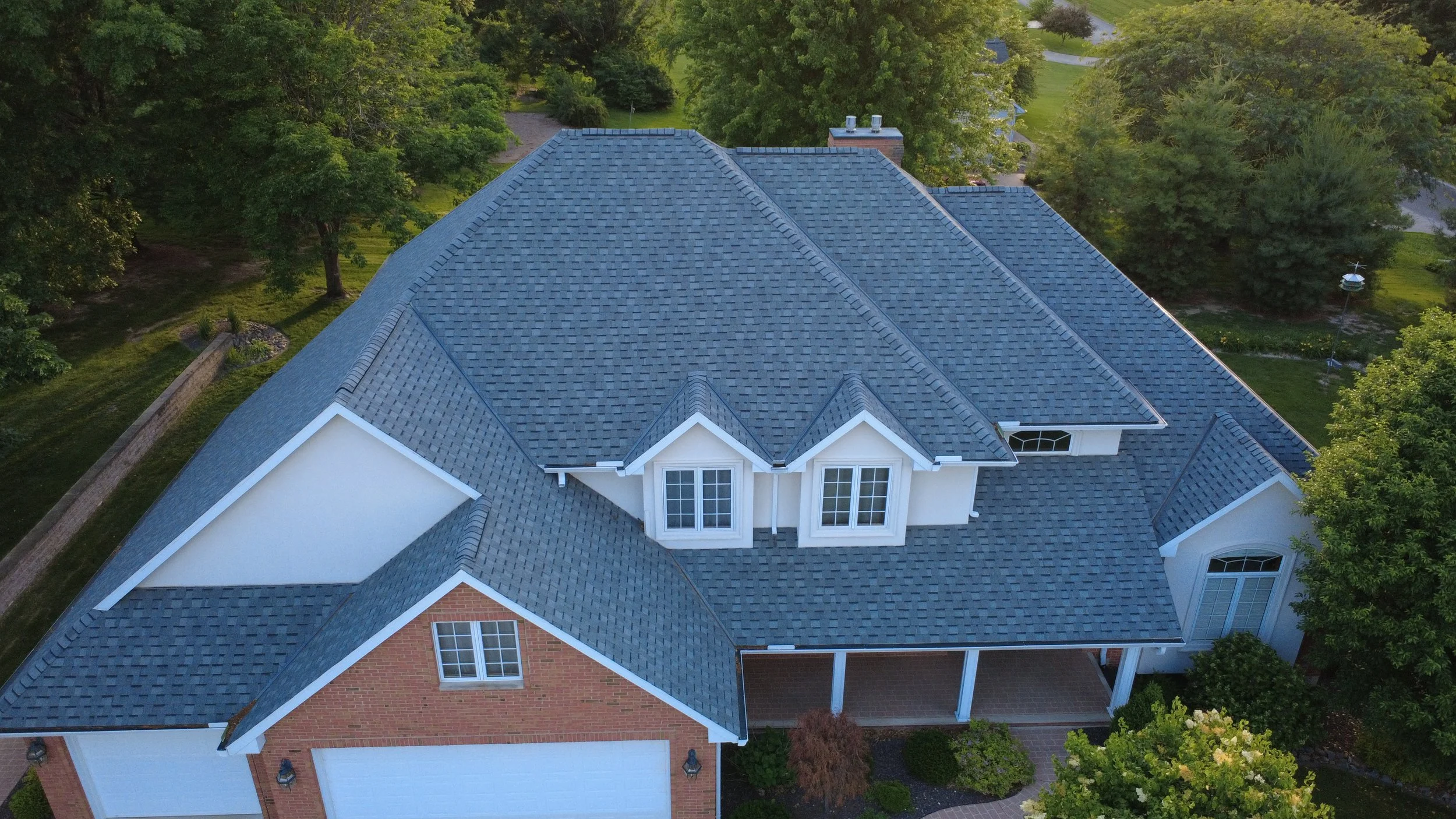 An aerial view of a house with a blue gabled roof, surrounded by green trees and a manicured lawn.