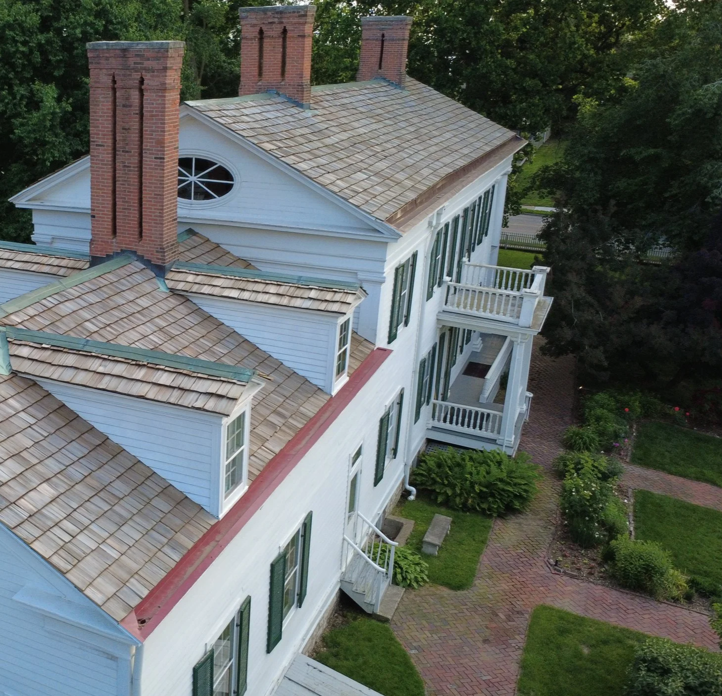 Aerial view of a large, historic white house with multiple brick chimneys, green shutters, and a porch surrounded by a garden with brick pathways and green foliage.