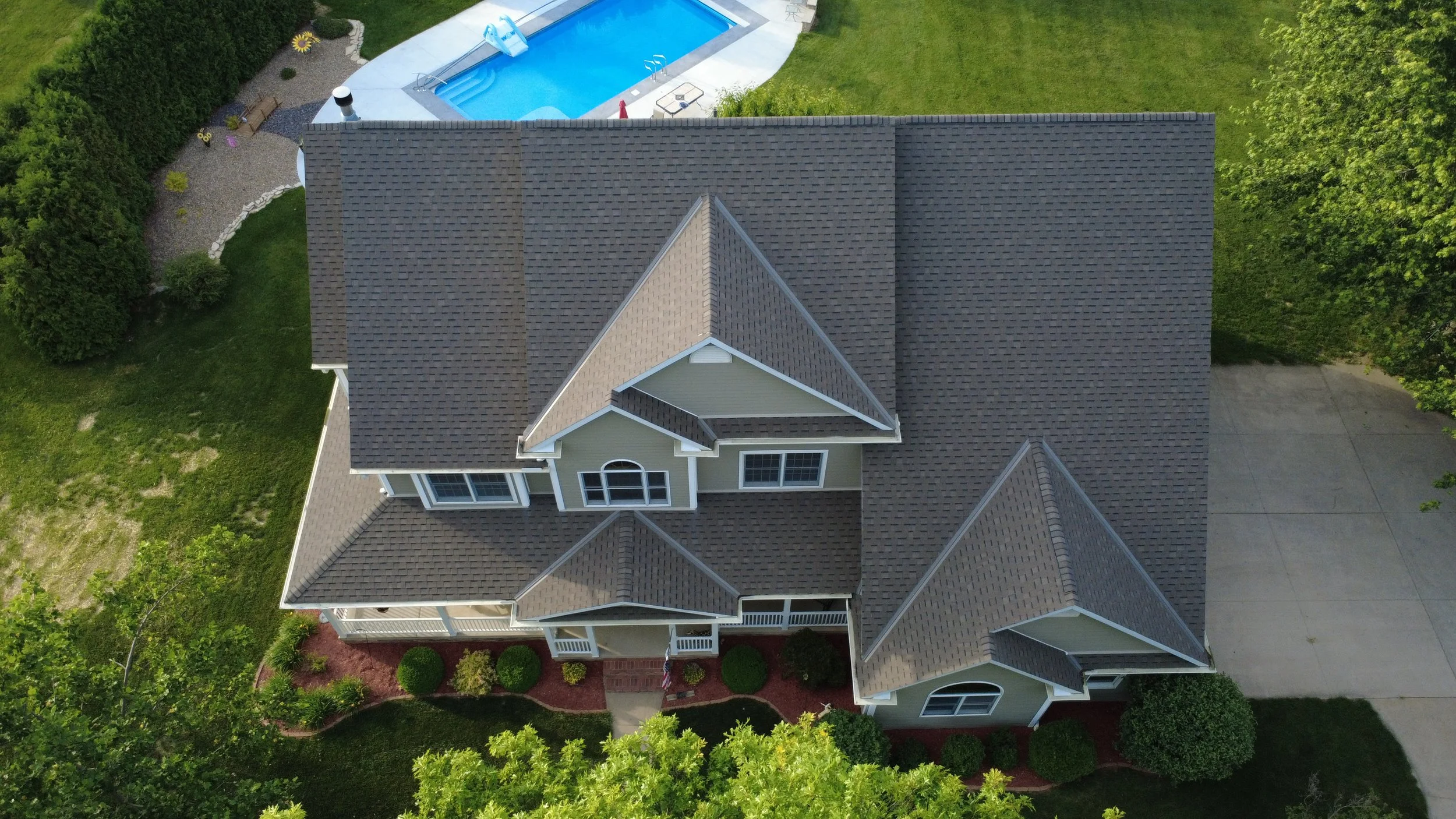 Aerial view of a house with a dark gray shingle roof, surrounded by green trees and grass. There is a backyard with a swimming pool and a small patio area.