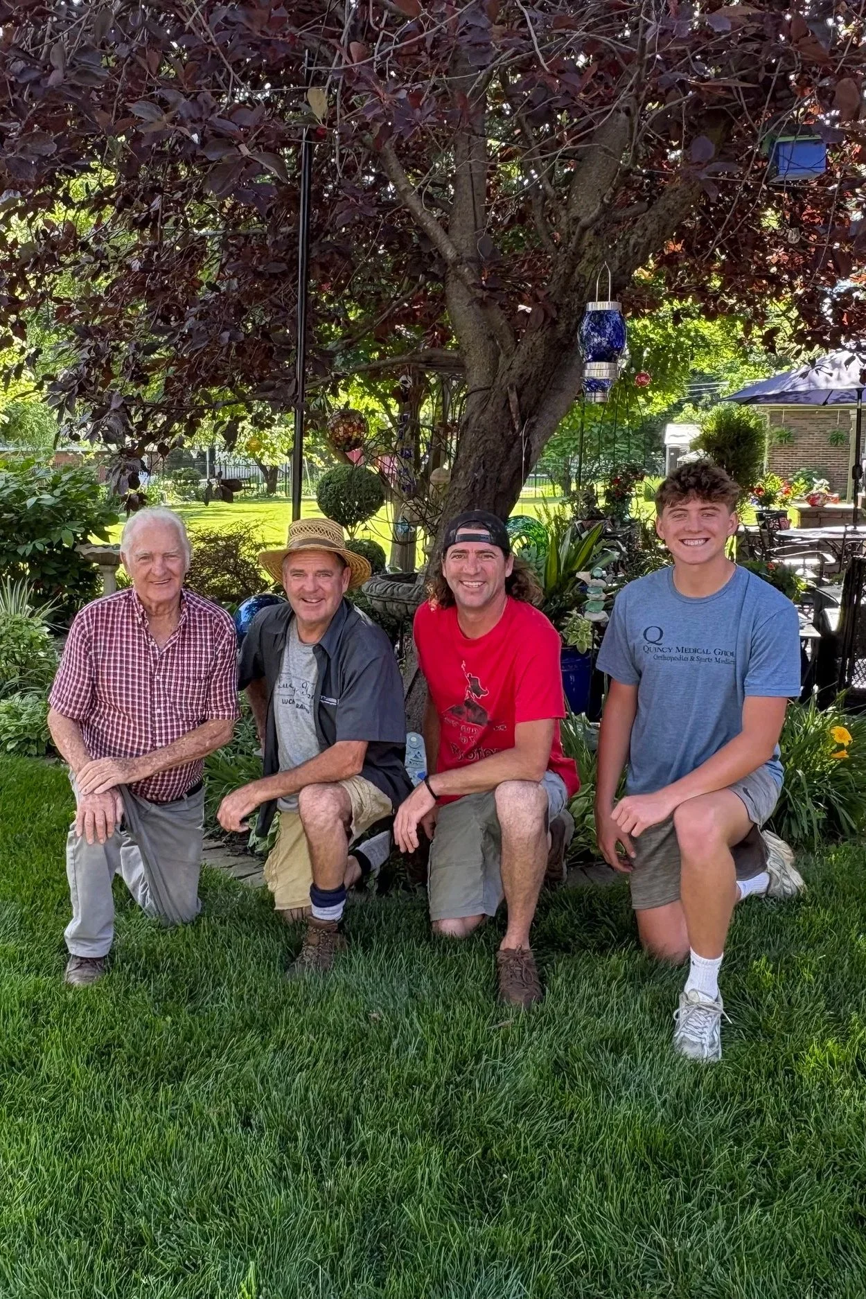 Four males kneeling and smiling in a backyard garden under a large tree. They are the 4 generations of the company.