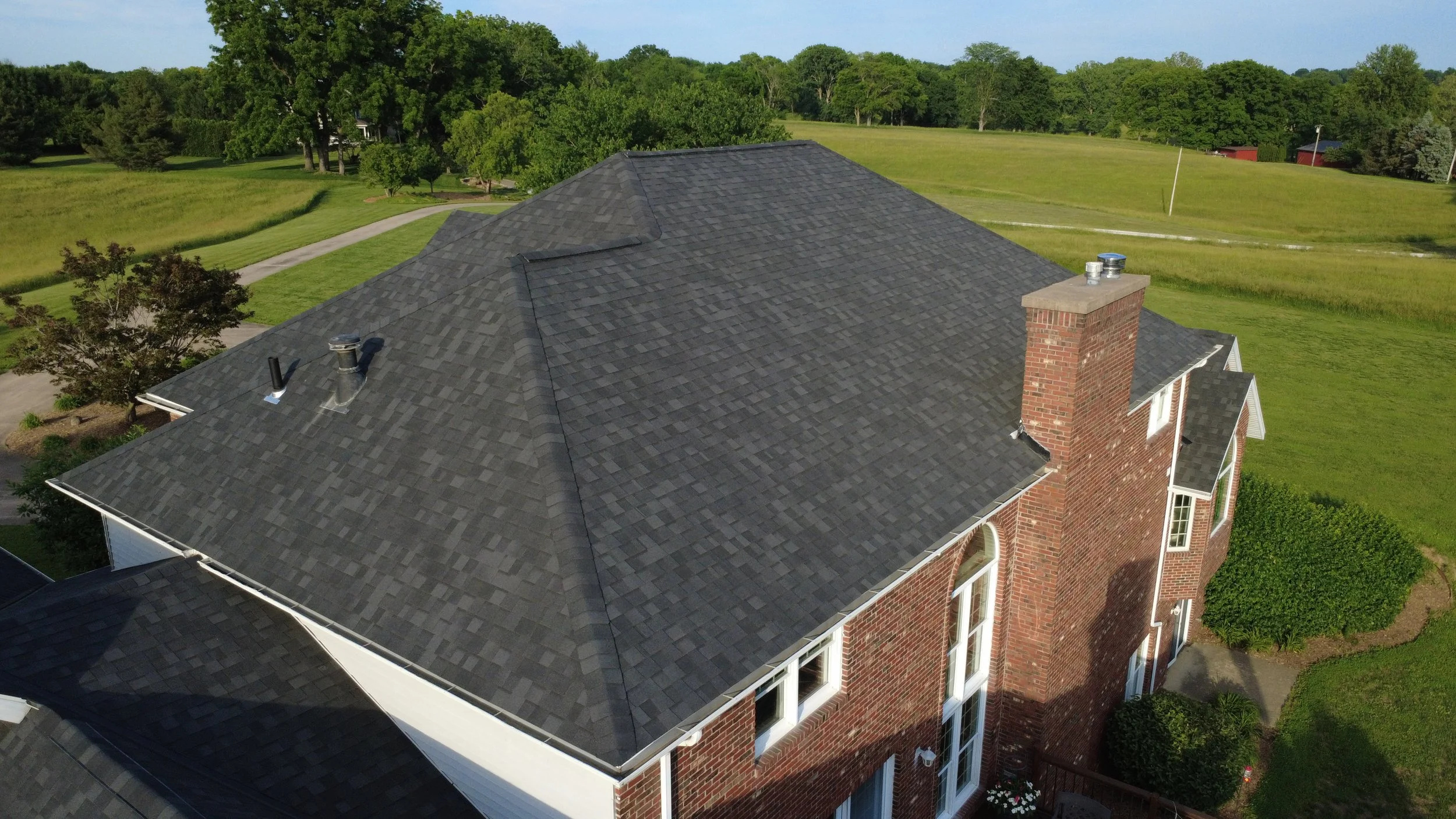 Aerial view of a brick house with a dark gray shingled roof, surrounded by green lawns, trees, and open fields with a pathway and utility pole in the background.