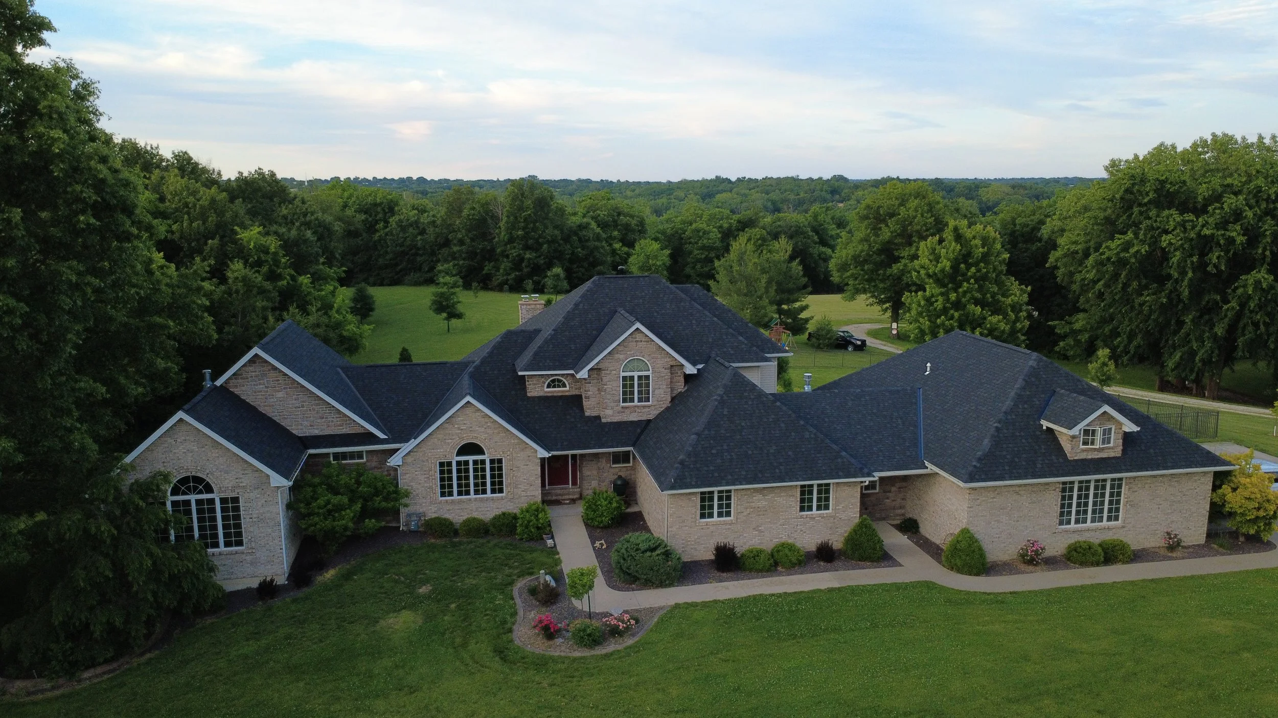 A large house with brick walls and dark shingle roof, surrounded by lush green grass and trees, with a spacious backyard and distant wooded area under a partly cloudy sky.