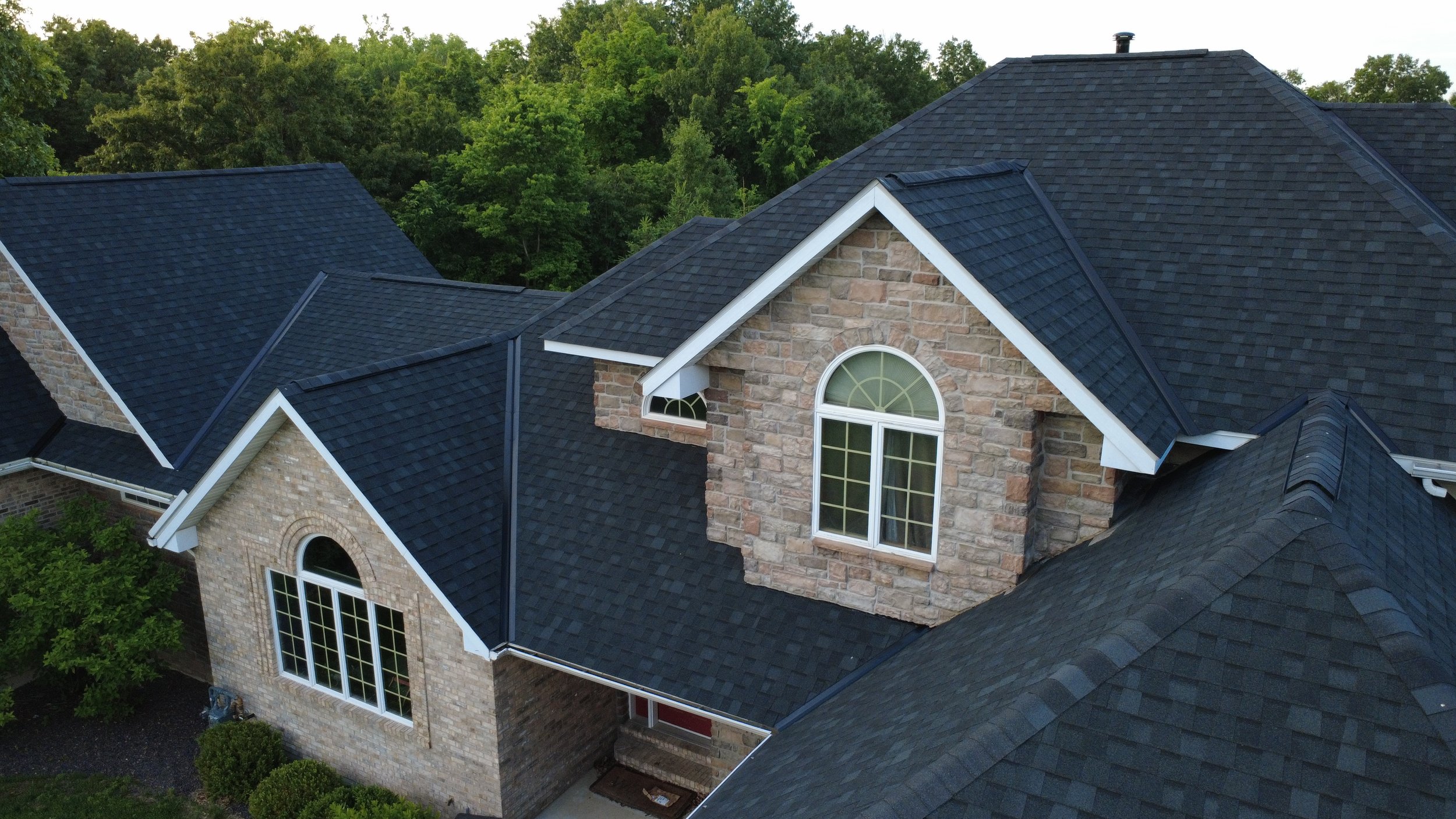 Aerial view of a house with a dark shingle roof, brick and stone exterior, arched windows, surrounded by green trees.