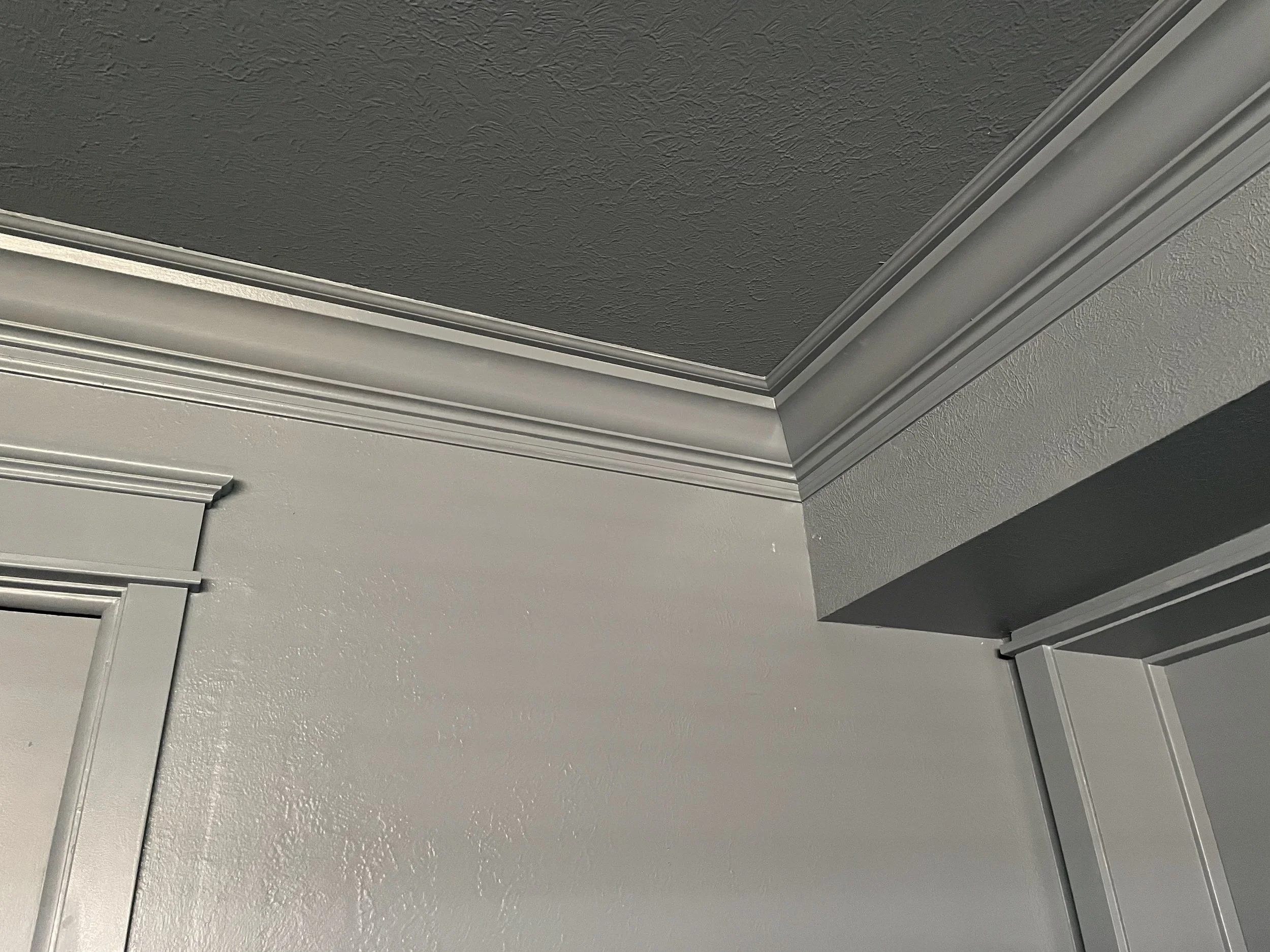 The image shows a close-up of the corner of a ceiling with crown molding. The ceiling is painted dark gray, and the walls are painted a lighter gray. The molding is painted in a light gray color.