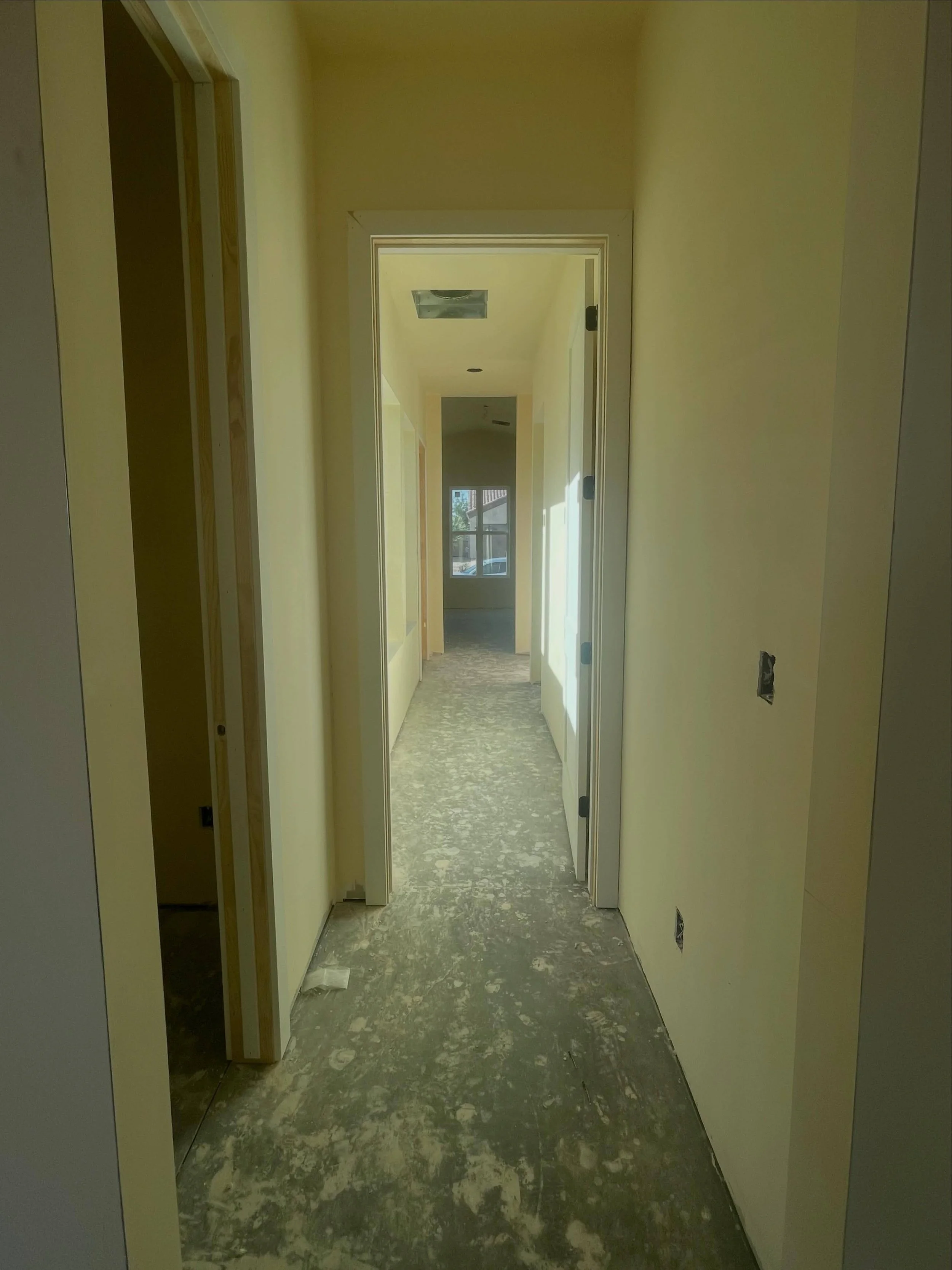 A narrow hallway in a house under construction or renovation, with unfinished flooring and light yellow painted walls. Doors on both sides and at the end, with a window at the far end letting in natural light.