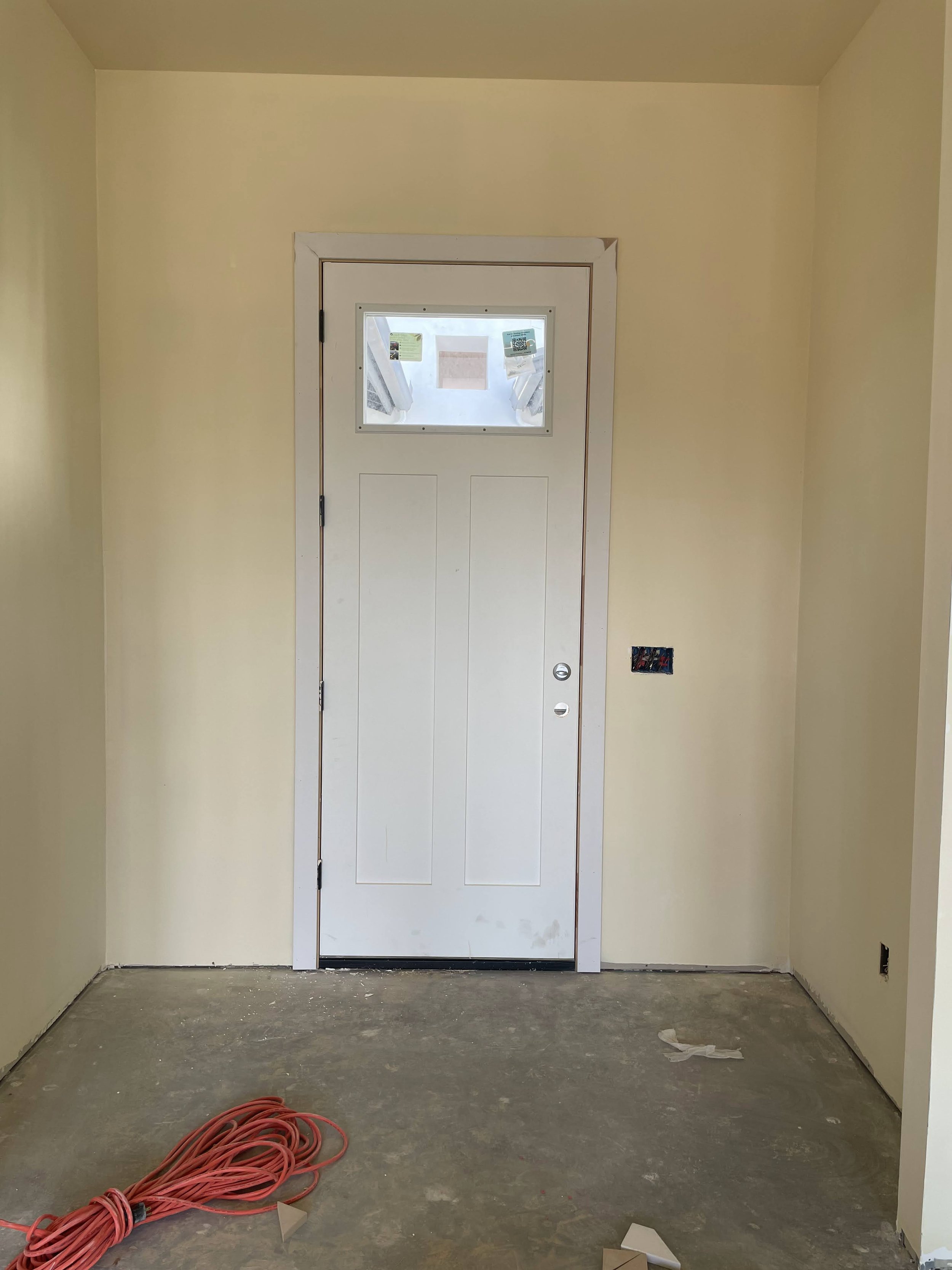 Interior of a room under construction with a white door and a small window, yellow walls, unfinished flooring, and red extension cord on the floor.