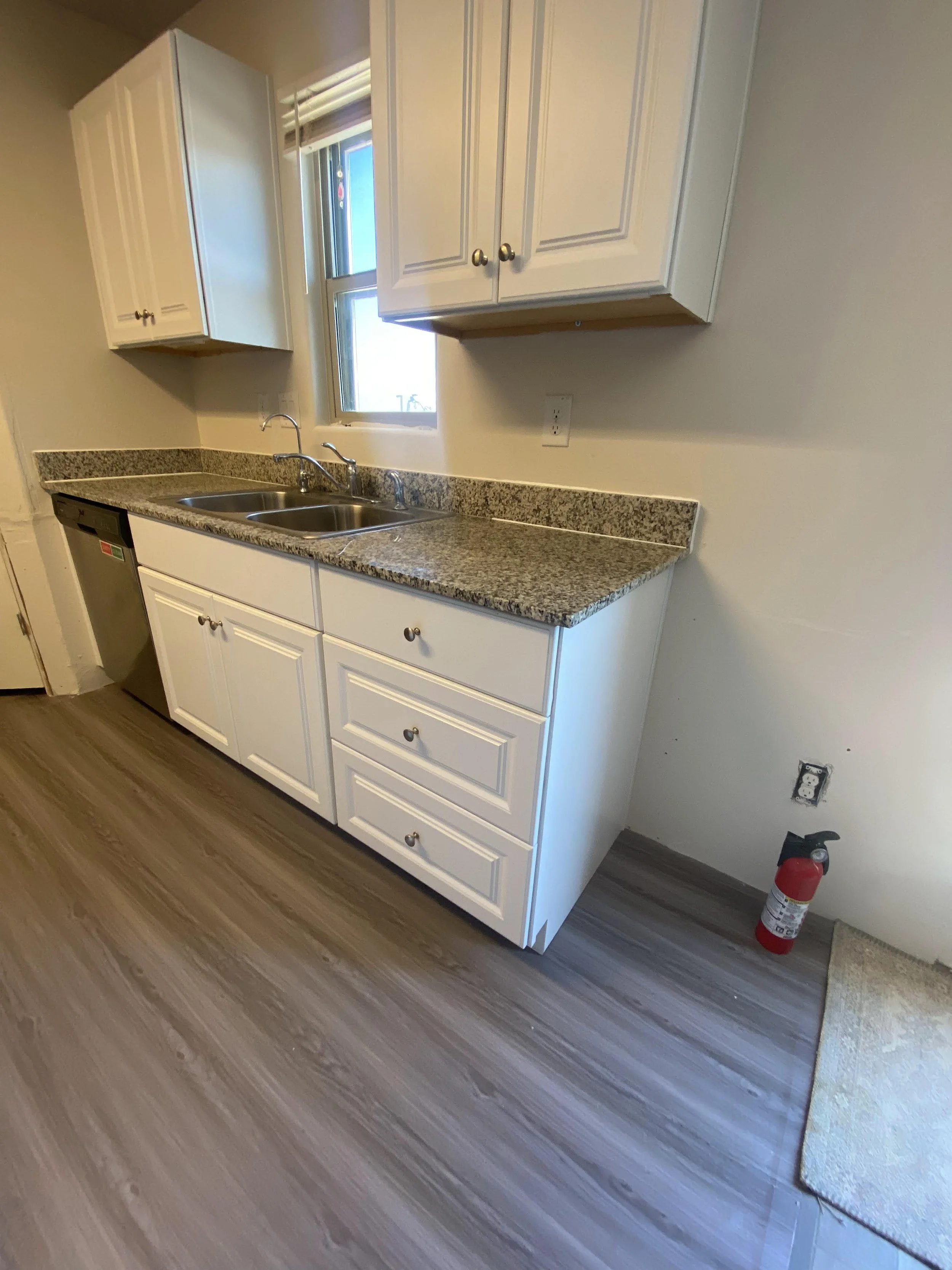 Kitchen with white cabinets, granite countertops, double sink, window above, and wood-look flooring. Fire extinguisher on the floor near wall outlet.