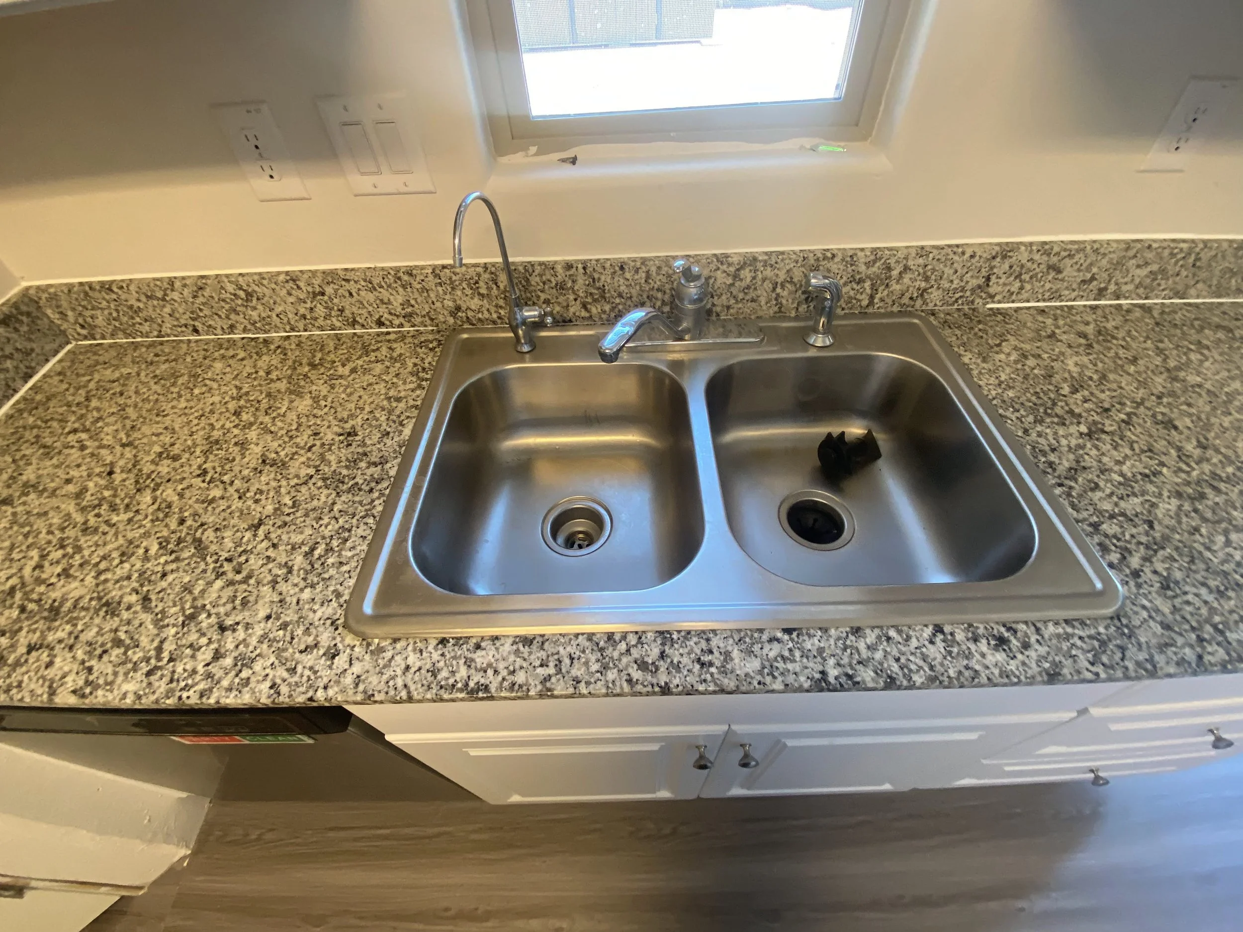 Kitchen countertop with a stainless steel double sink, granite surface, and a window above the sink. There are electrical outlets on the wall above the counter.