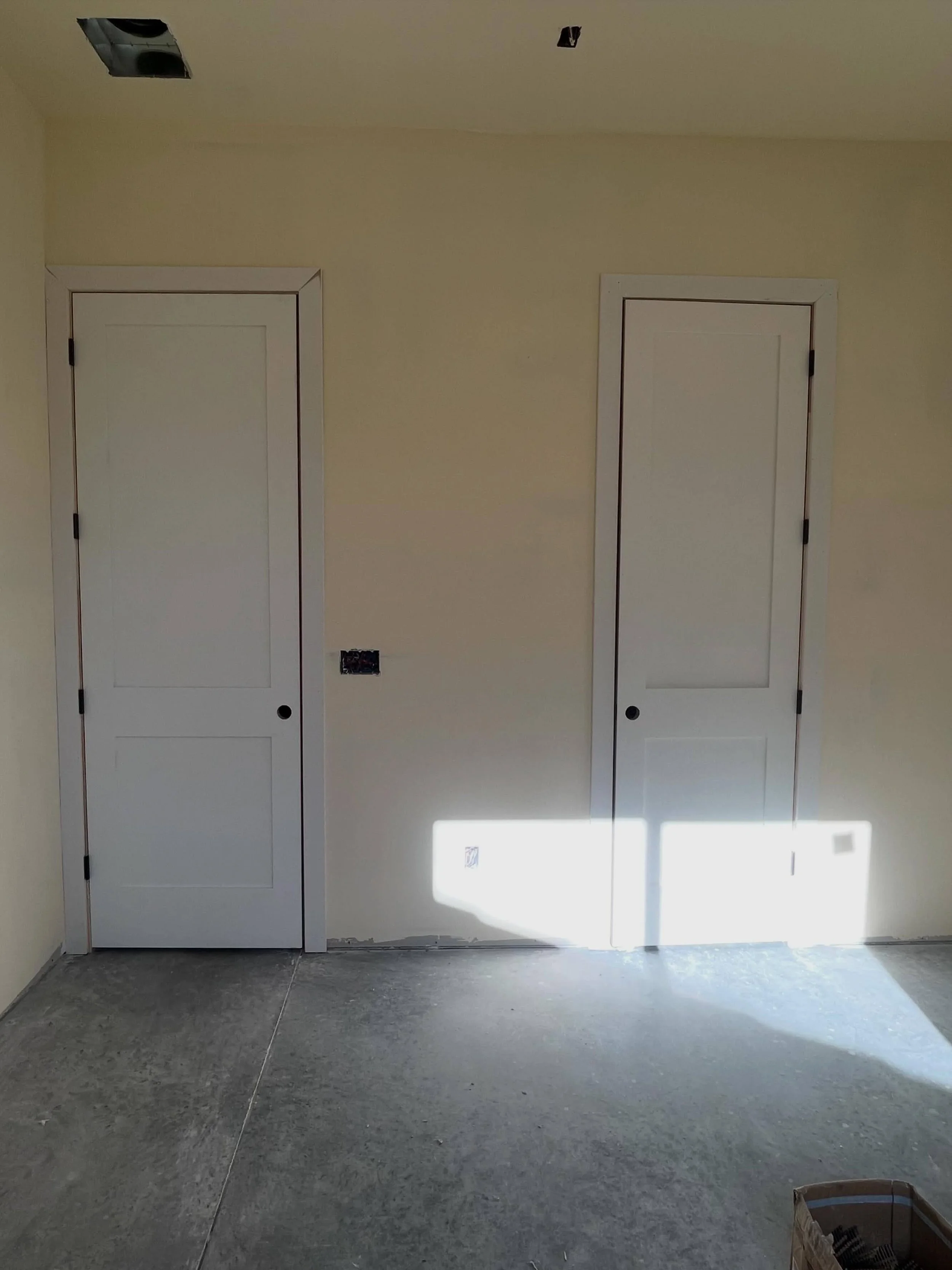 Two white doors with black hinges and door knobs in a beige wall. There are two open ceiling panels above the doors, and an electrical outlet at the bottom of the wall. The floor is concrete, and a cardboard box with black objects is on the bottom ri