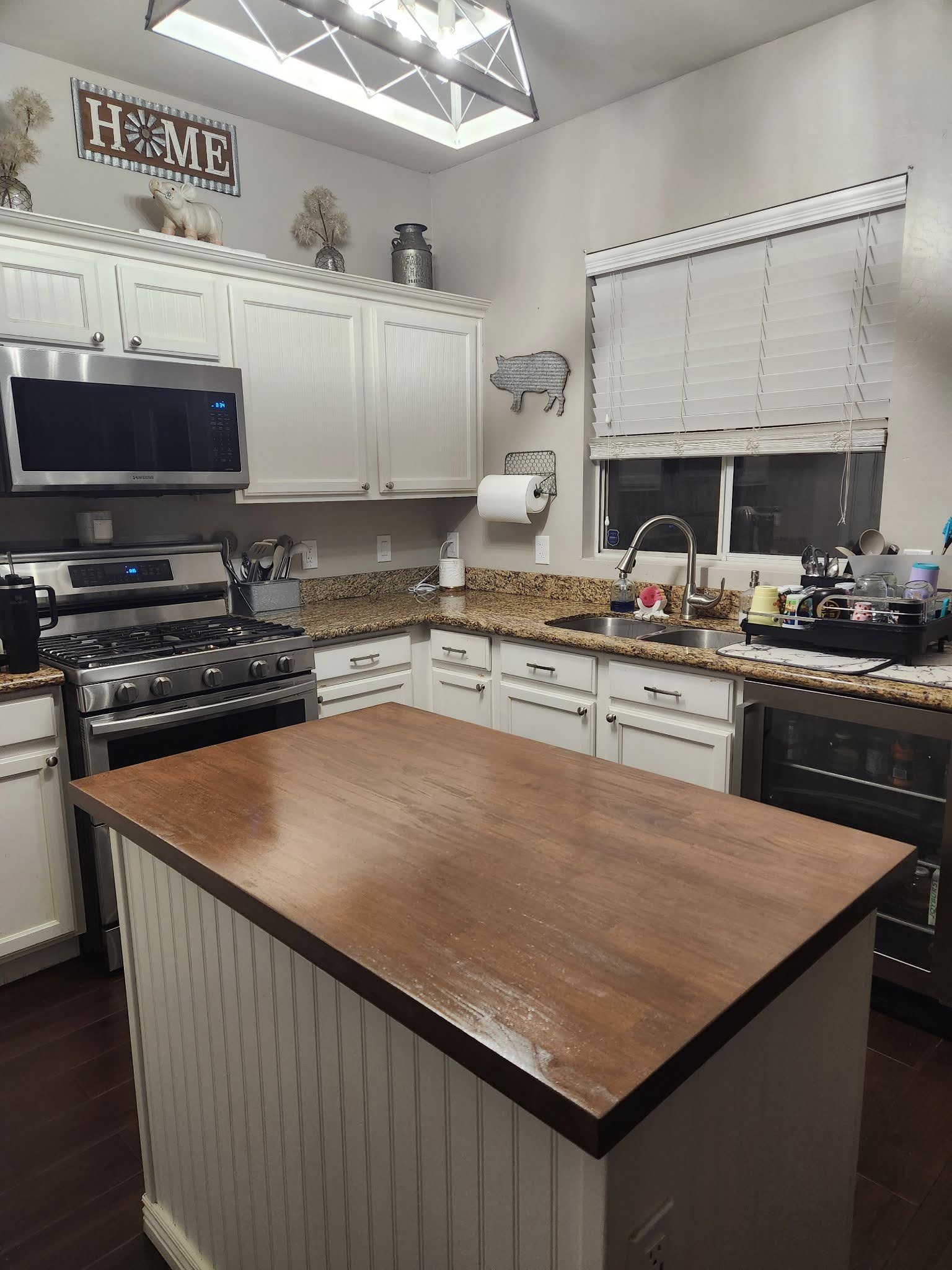 A kitchen with white cabinets, a granite countertop, a stainless steel microwave and oven, a wooden kitchen island, a window with white blinds, and decorative items including a 'HOME' sign, a pig figurine, and plants on top of the cabinets.
