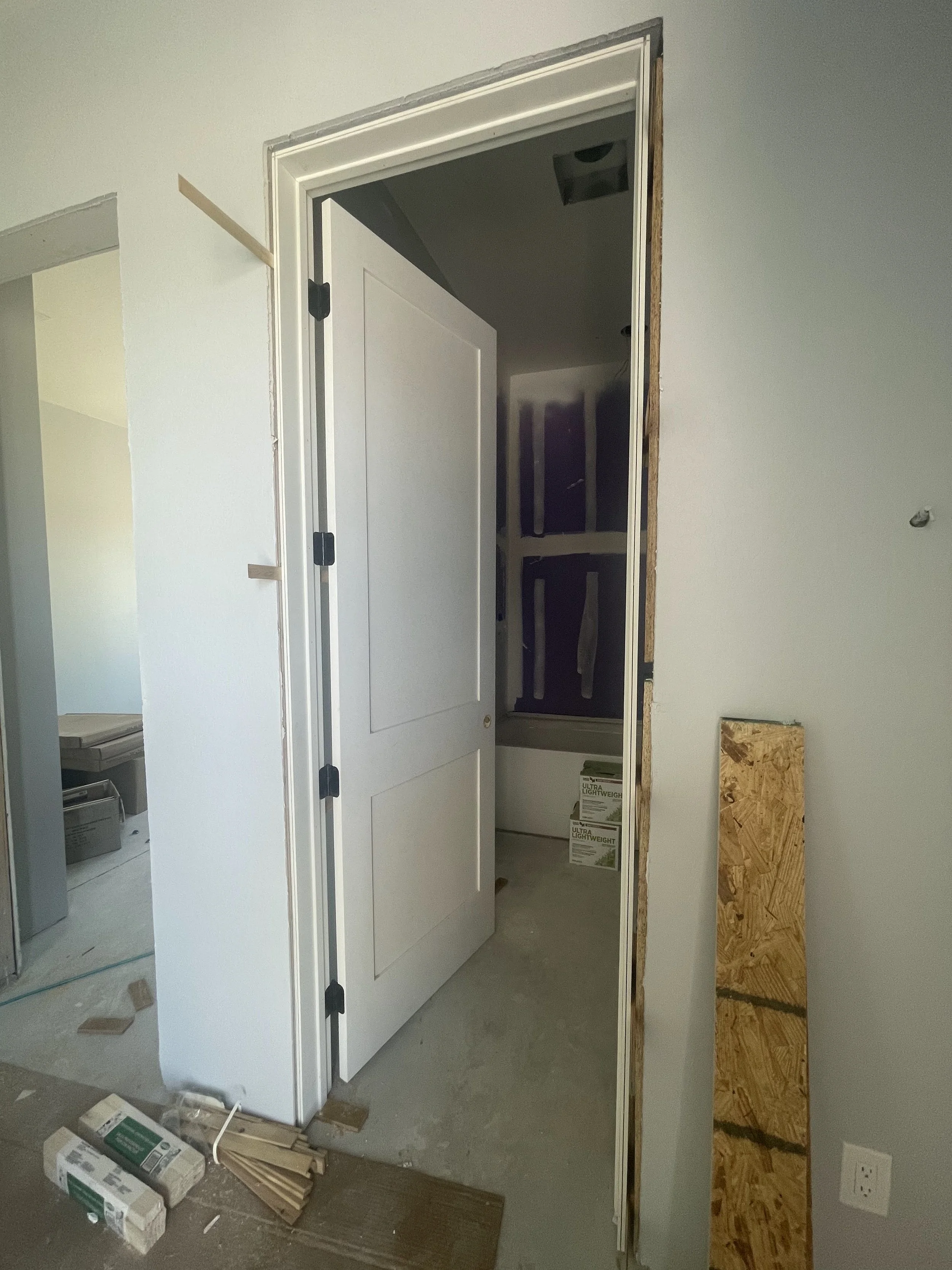 Interior of a house under construction showing an shaker style door installation with black hinges, drywall, and unfinished walls, with construction materials on the floor.