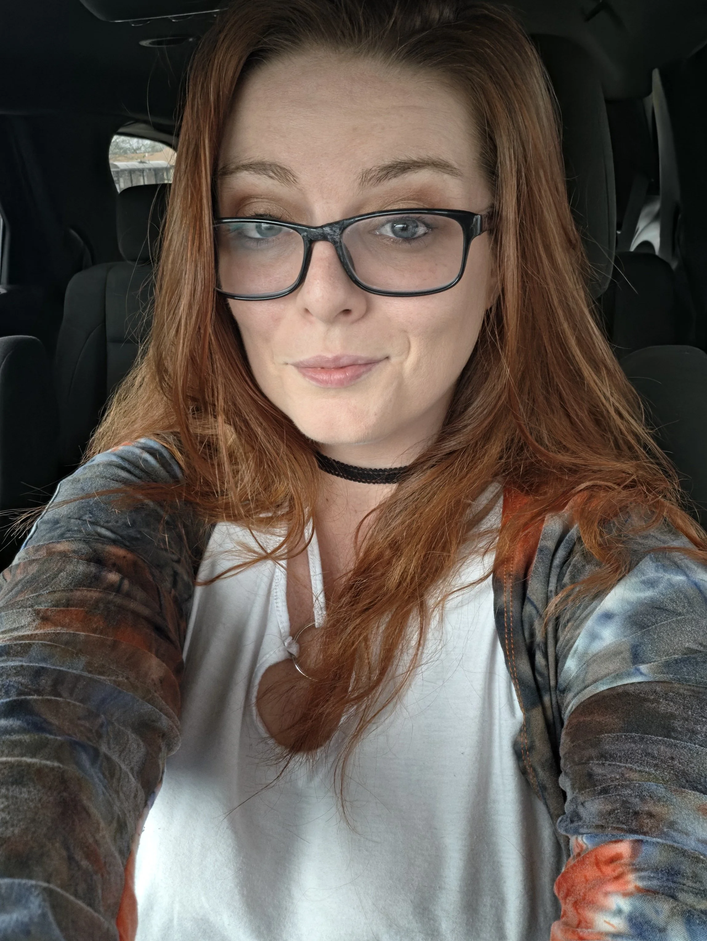 Close-up of a woman with red hair and glasses, smiling inside a vehicle.