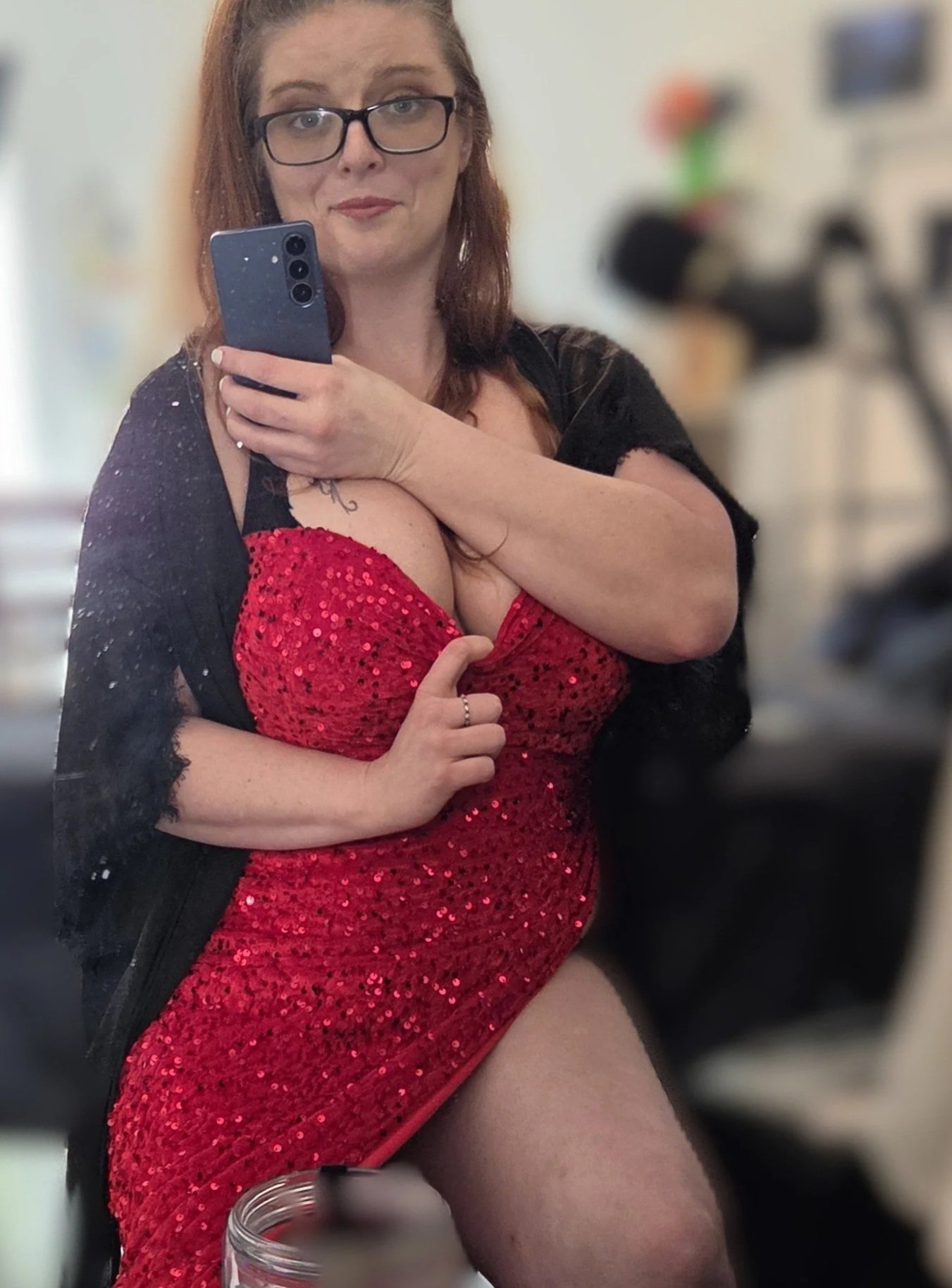 Woman wearing glasses and a red sequined dress takes a mirror selfie with a smartphone.