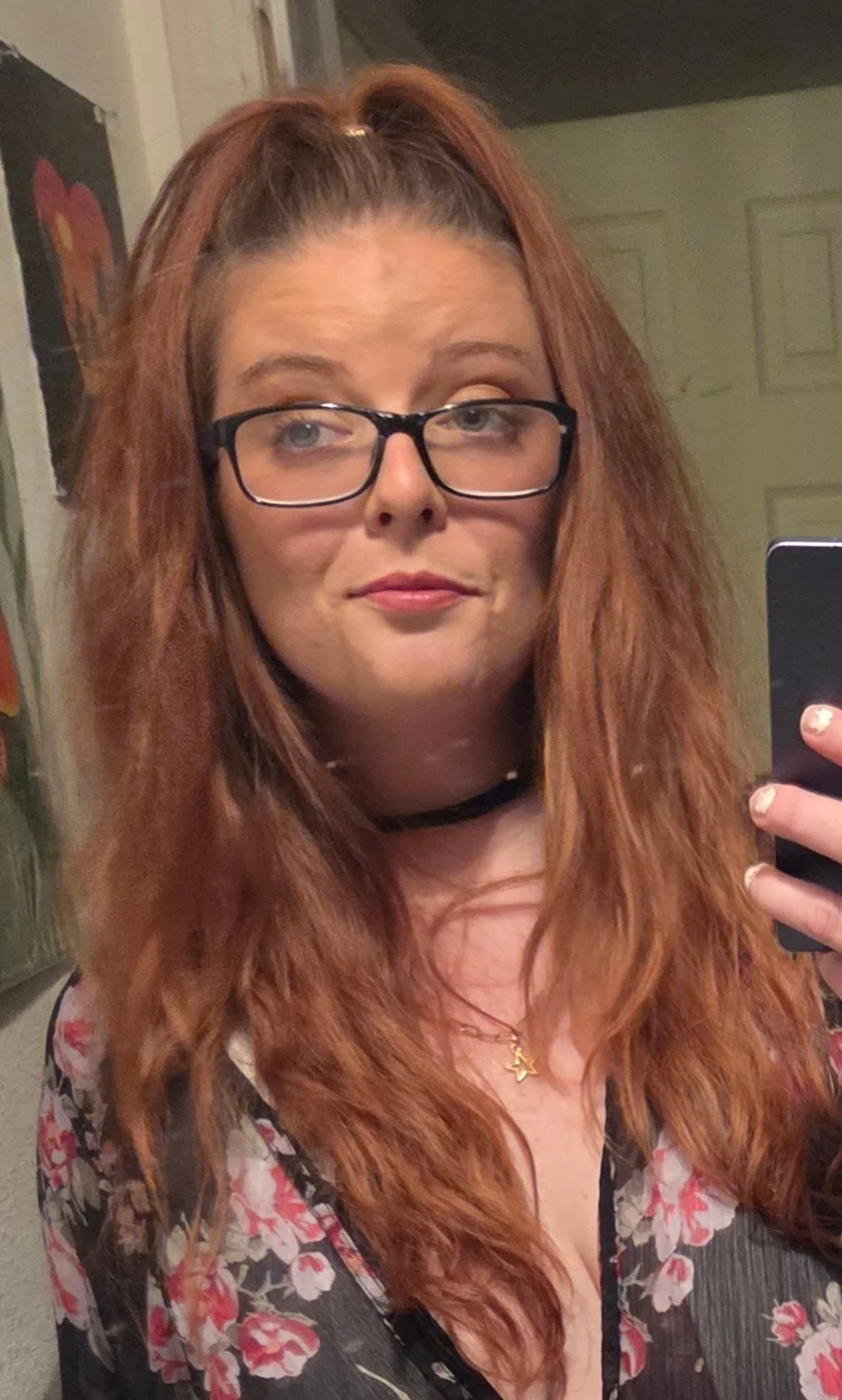 Woman with long auburn hair and glasses taking a mirror selfie indoors