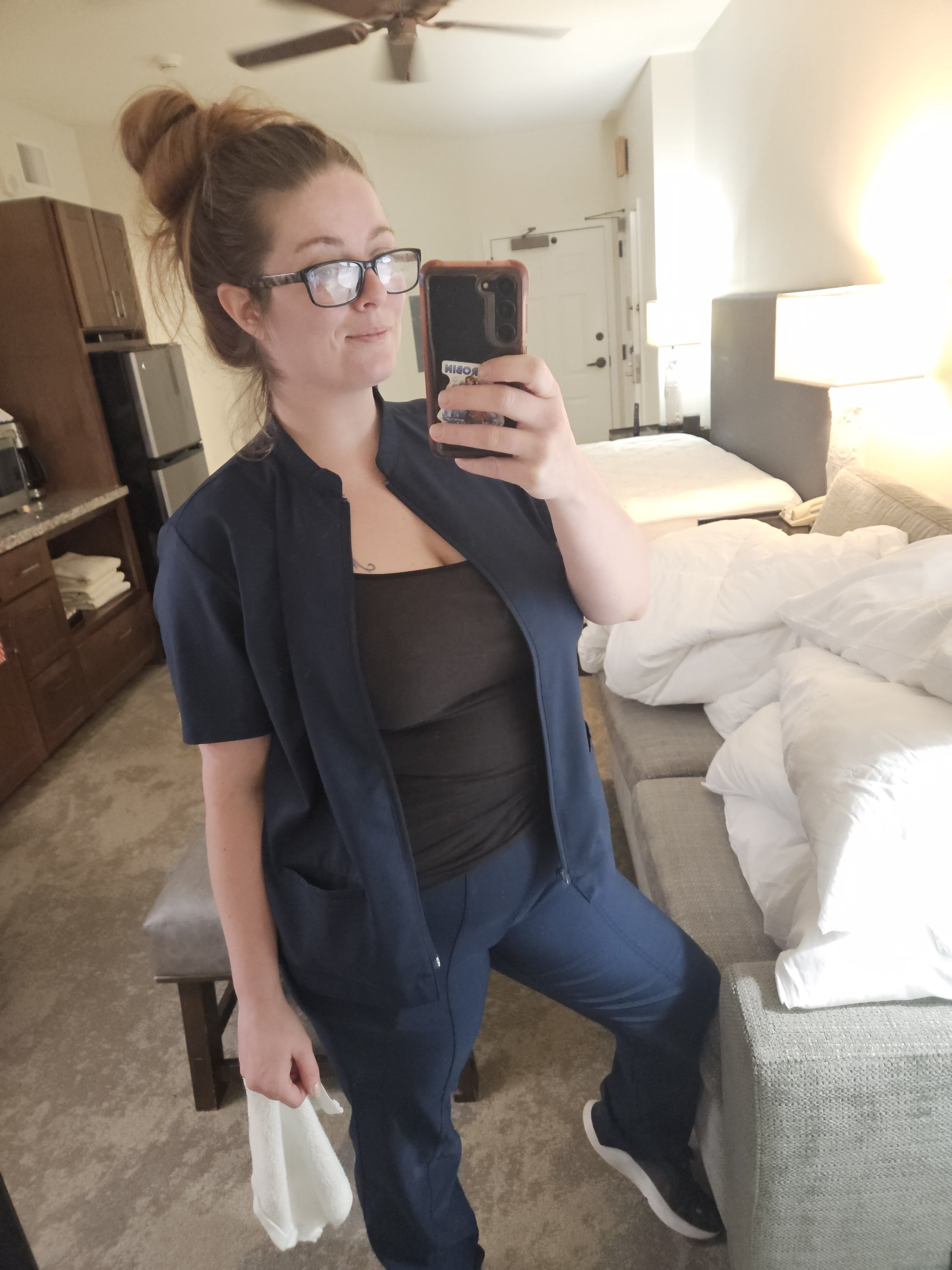 A woman with glasses and a bun hairstyle taking a selfie in a hotel room mirror, with a bed, nightstand, lamp, and other furniture visible in the background.