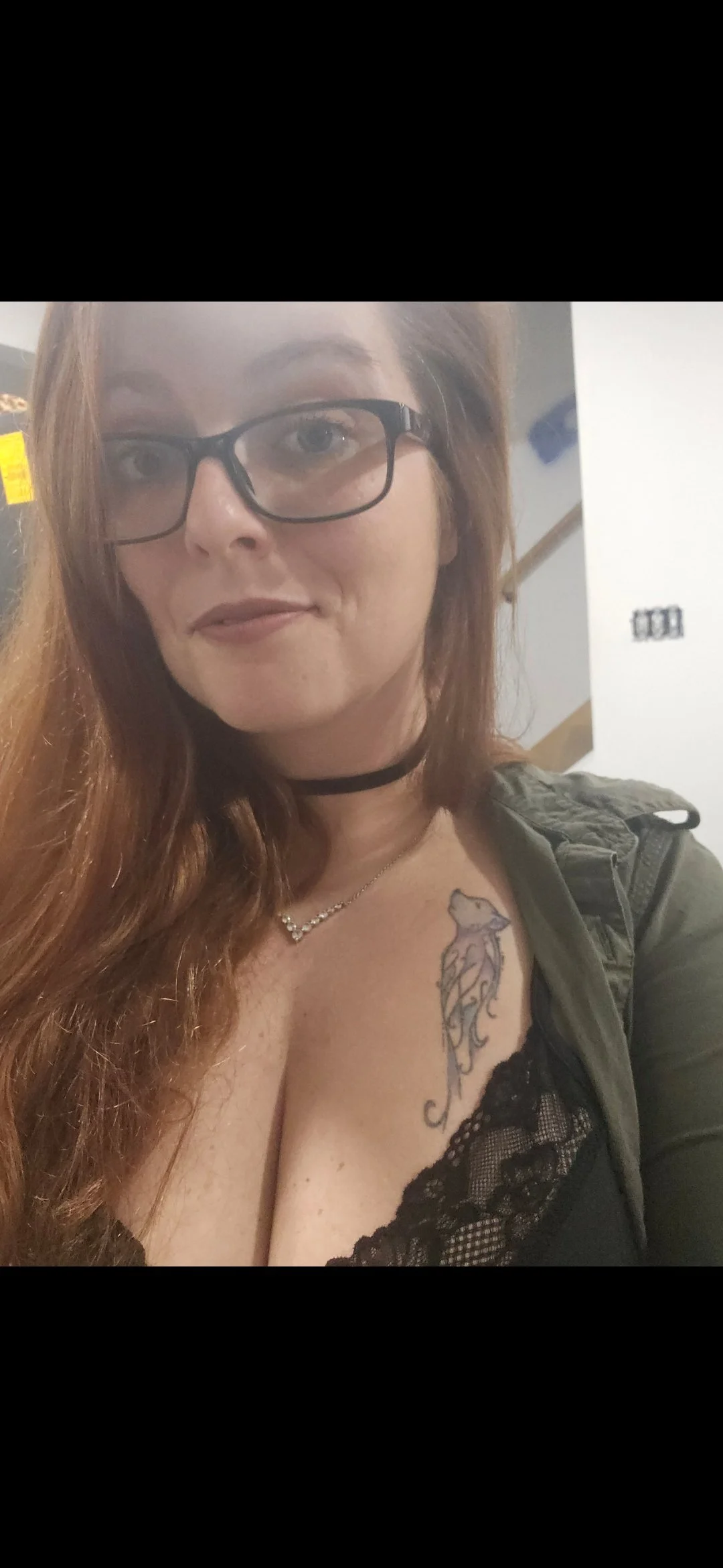 A woman with red hair, glasses, and a choker necklace taking a selfie, wearing a black lace top and a tattoo of a horse on her chest.