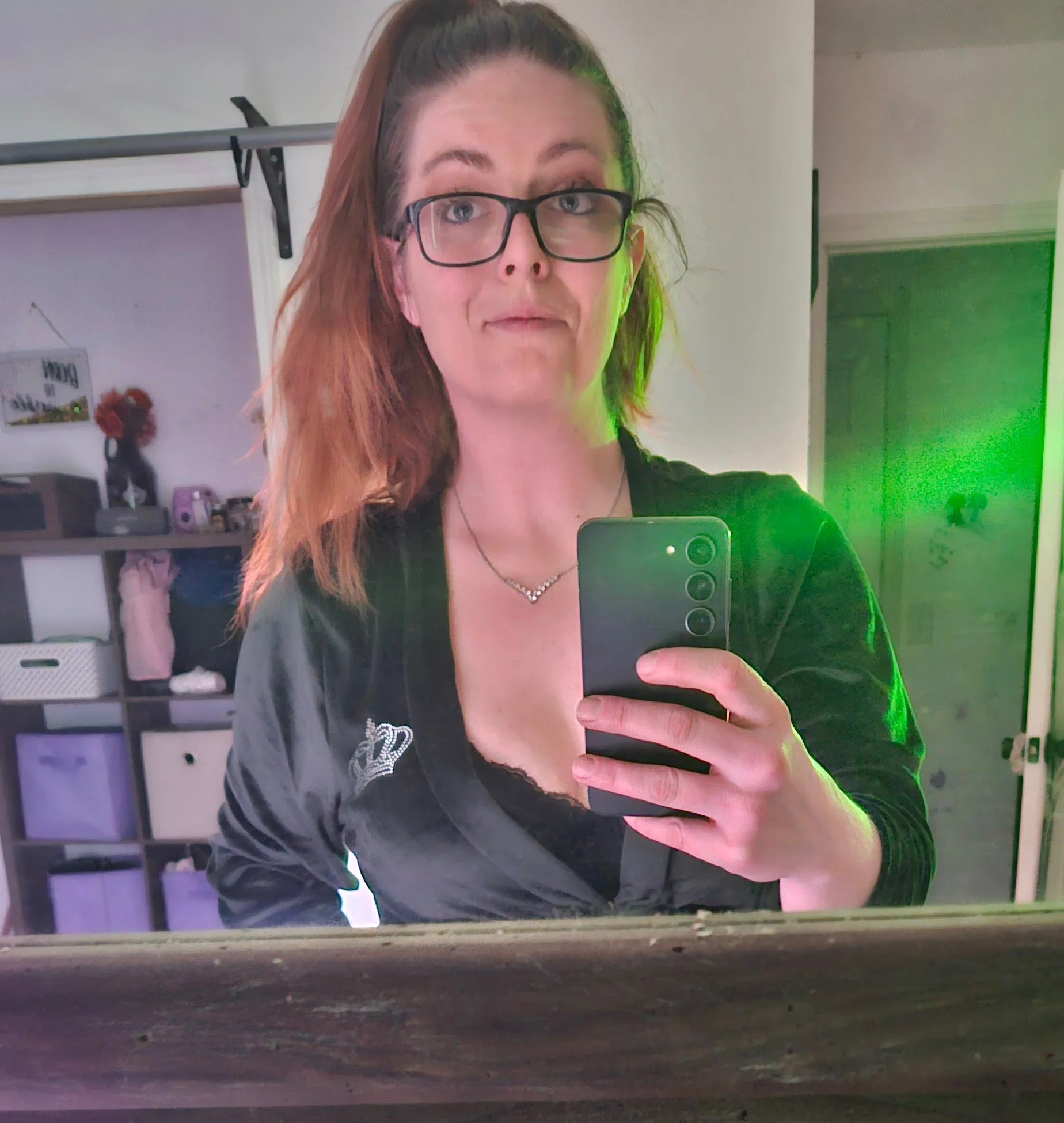 A woman with red hair and glasses taking a selfie in front of a mirror, holding a smartphone, with a background that includes shelves, a small statue, and various objects in a room with light-colored walls.