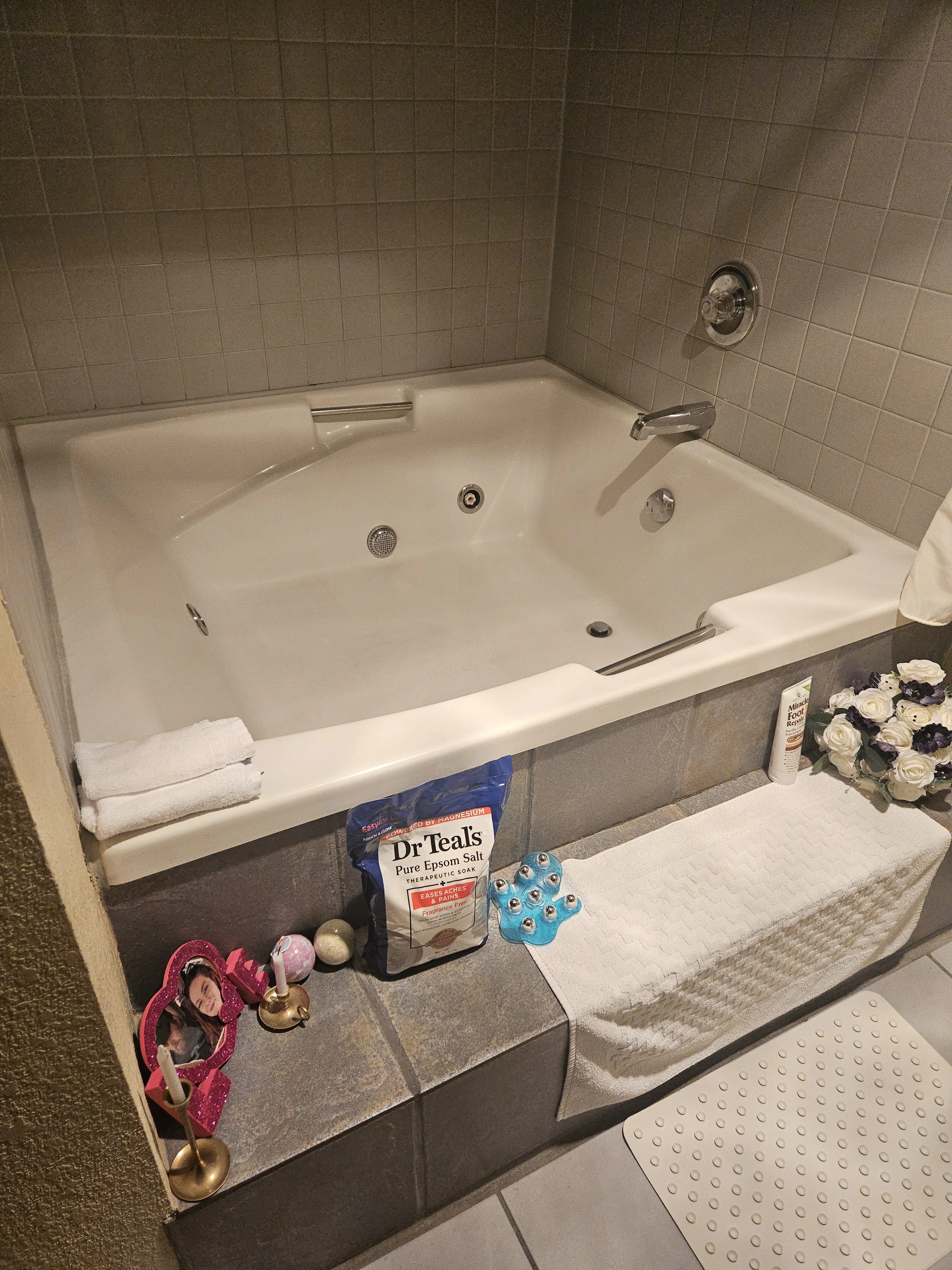Bathroom scene with a bathtub, towels, a bag of Epsom salt, decorative flowers, a pink picture frame, and some small colored objects on a tray. There's a white bath mat on the floor near the tub.