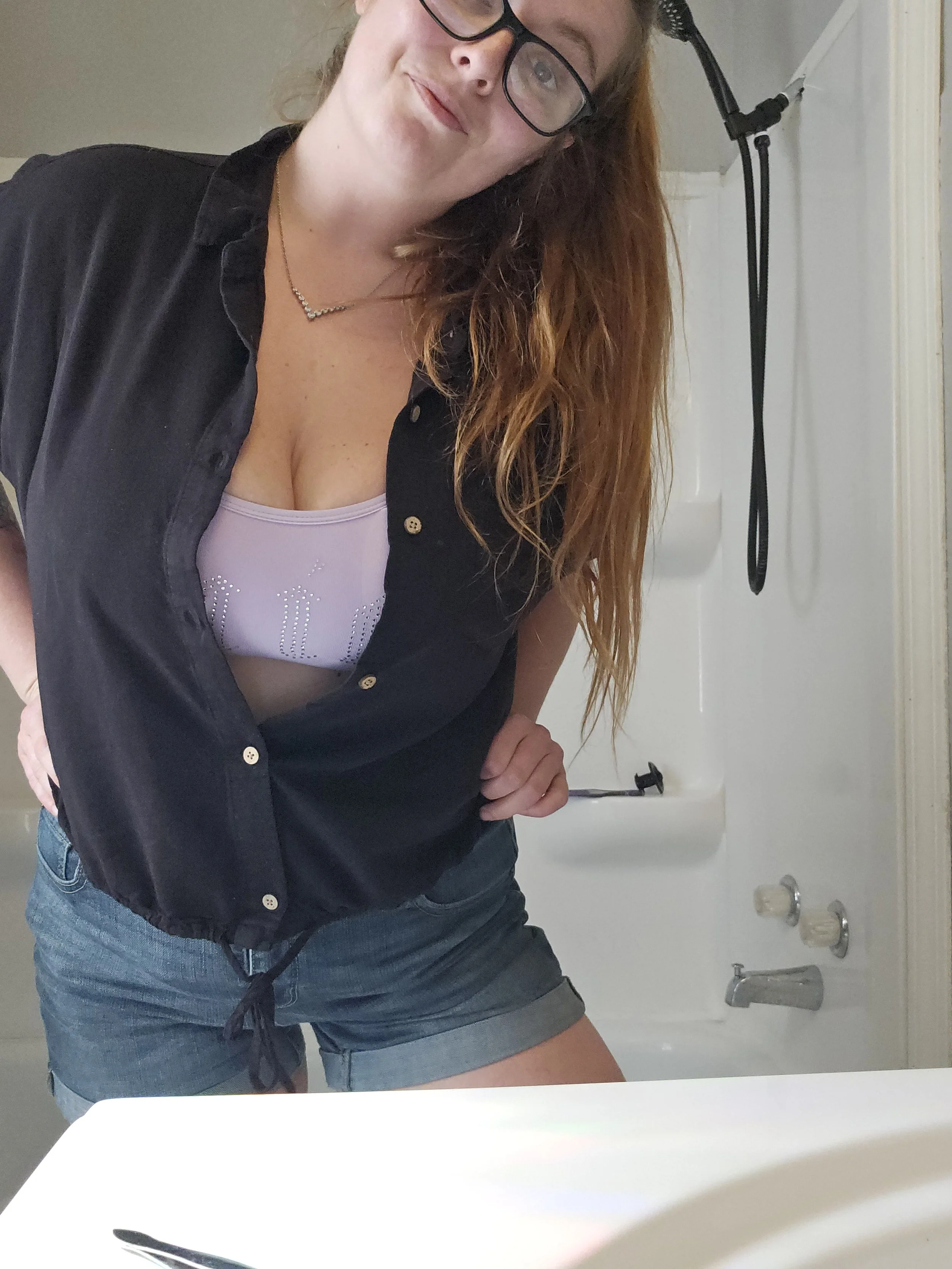 A woman is taking a mirror selfie in a bathroom. She has long red hair, wears glasses, a black shirt over a lavender tank top, and denim shorts. The bathroom features a white sink, a towel rack, and bathroom fixtures.