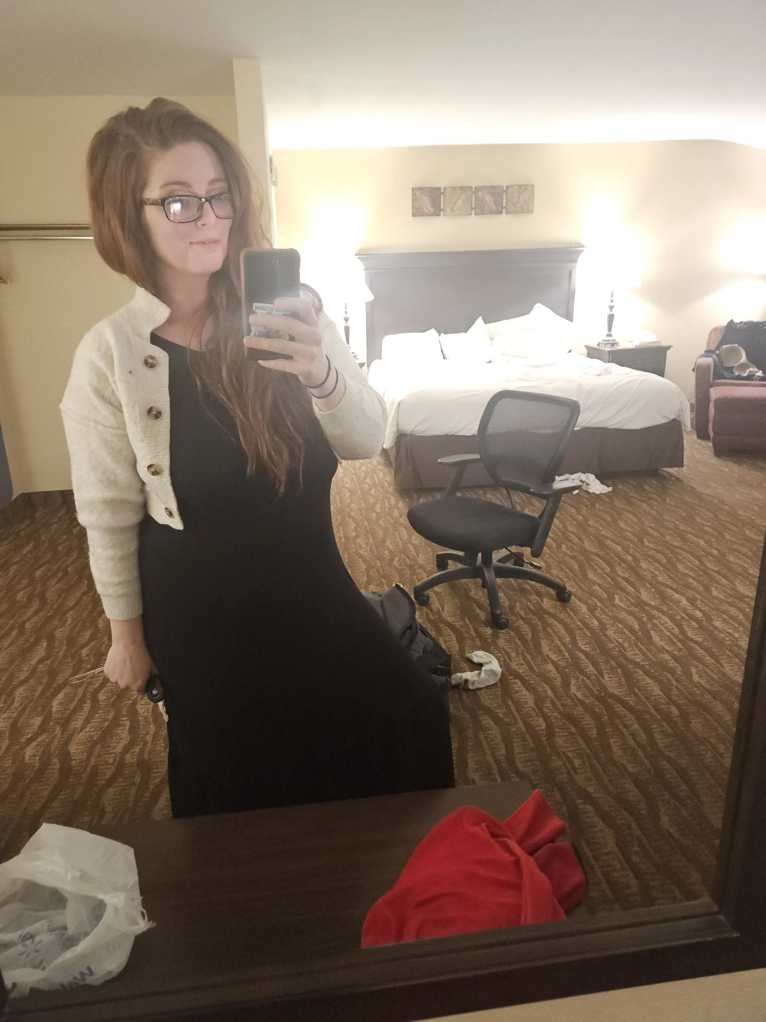 A woman with glasses and long hair taking a selfie in a mirror of a hotel room. The room has a bed with white linens and pillows, two nightstands with lamps, a chair, and a desk. A red hoodie and a white shopping bag are on the desk, and clothes are 
