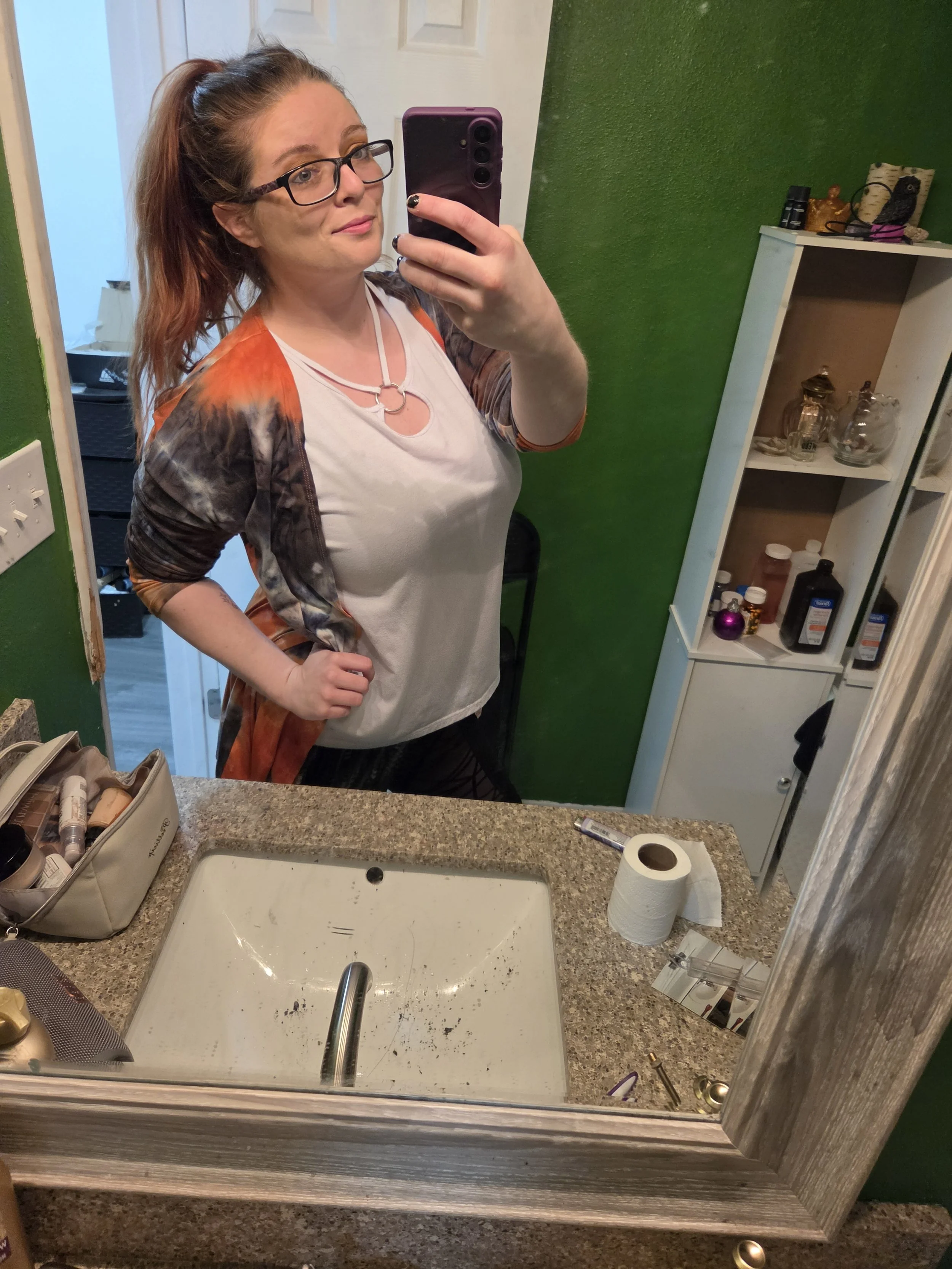 A woman with glasses taking a mirror selfie in a bathroom, with a green wall, white shelving unit, and a countertop with various items. The bathroom sink is visible and the countertop has makeup items, tissue paper, and other toiletries.