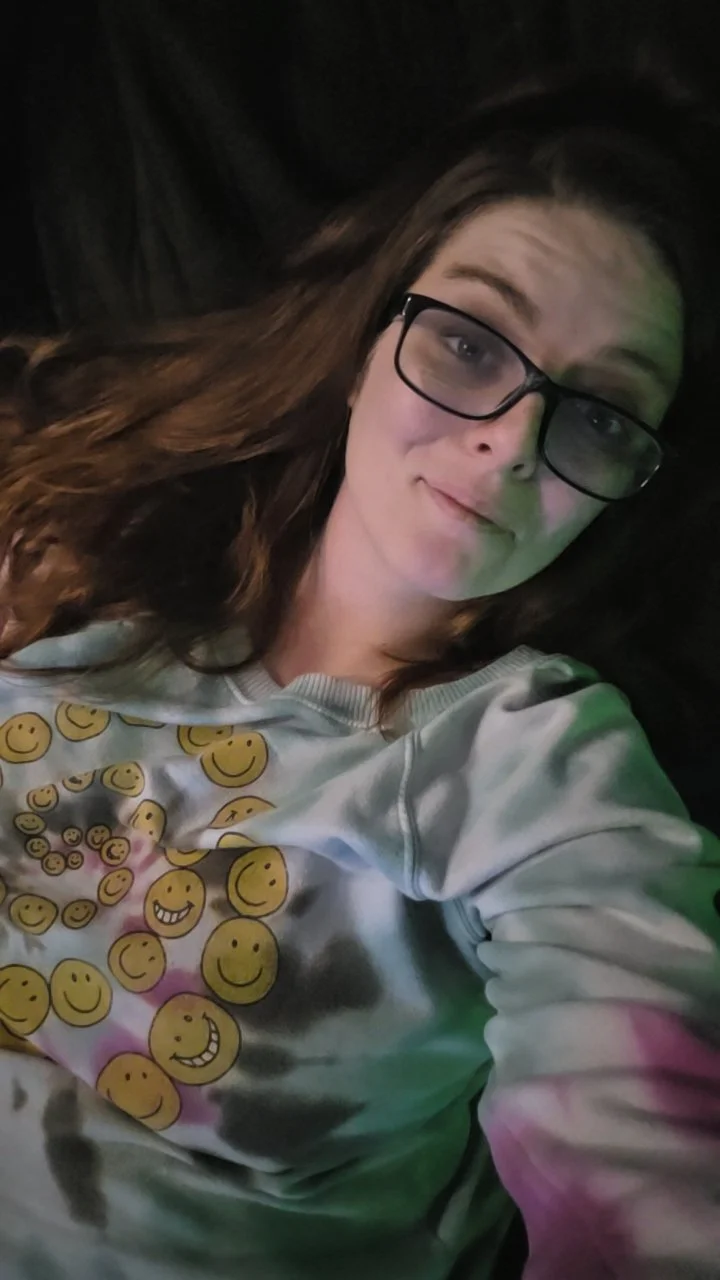 A young woman with long brown hair and glasses taking a selfie. She is wearing a white sweatshirt with various yellow smiley faces on it. The background is dark.