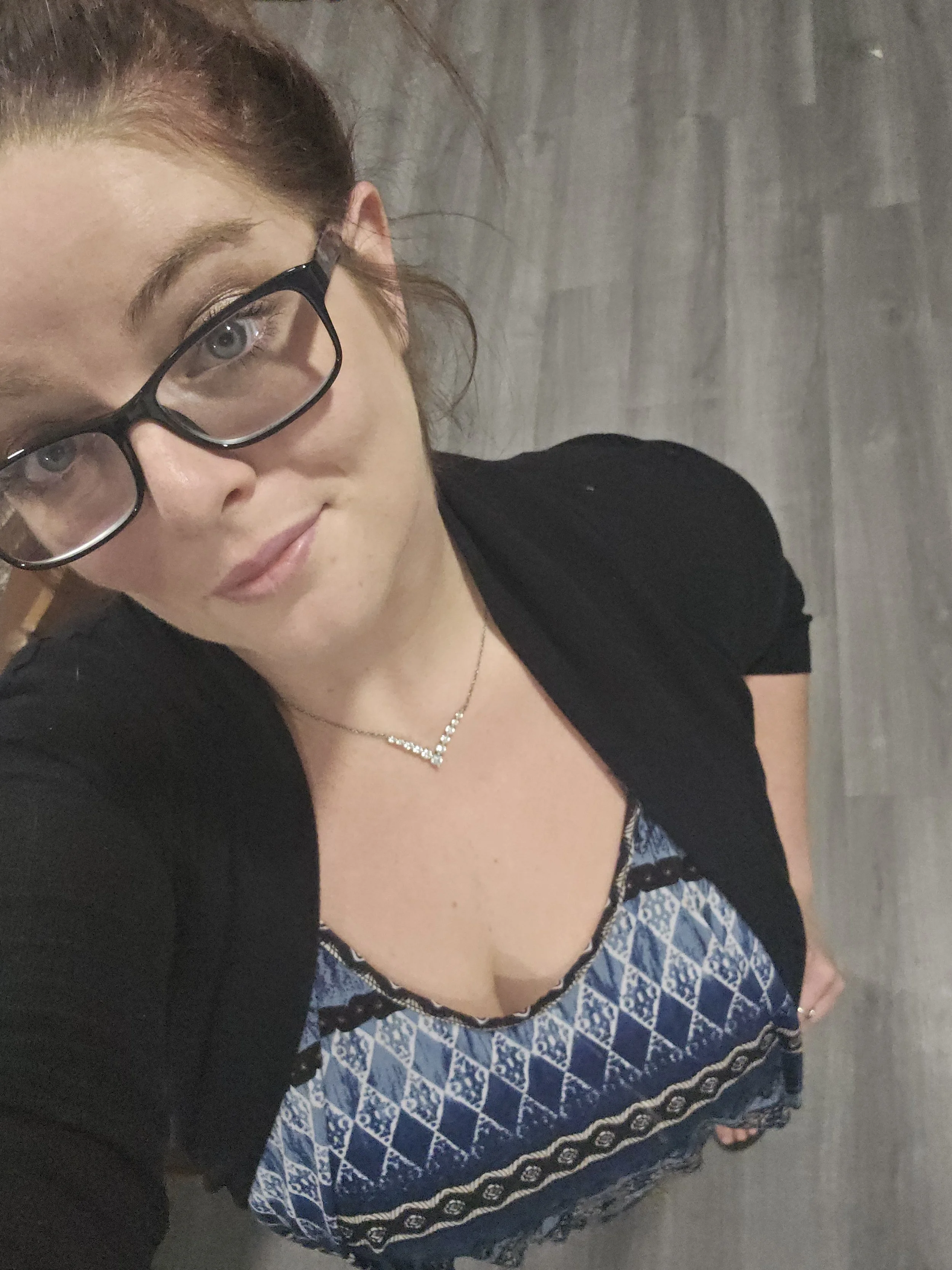 A woman with red hair and glasses wearing a black top and blue patterned skirt, standing on a gray floor.