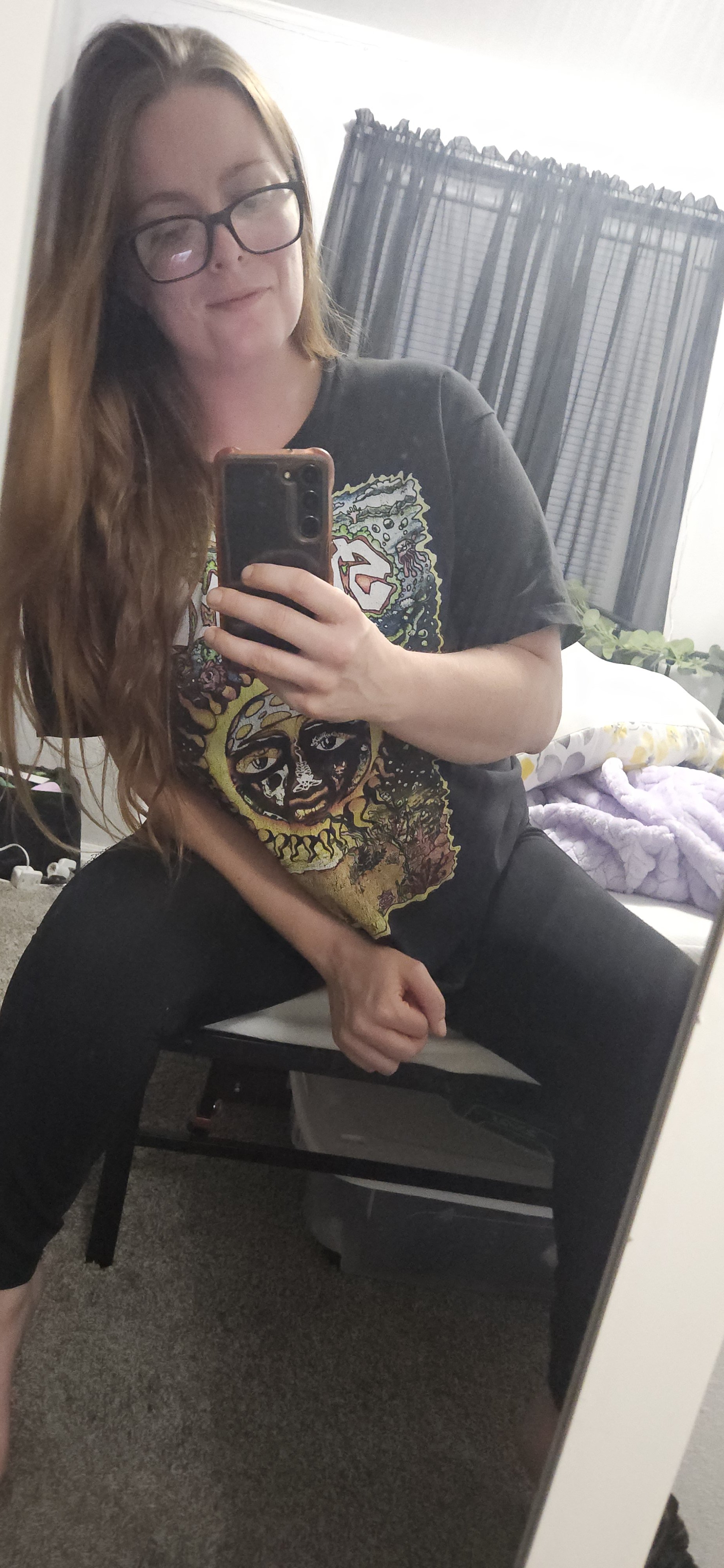 A woman with long hair and glasses taking a selfie in a mirror, wearing a black band t-shirt with colorful design, sitting on a chair in a room with dark curtains and a bed in the background.