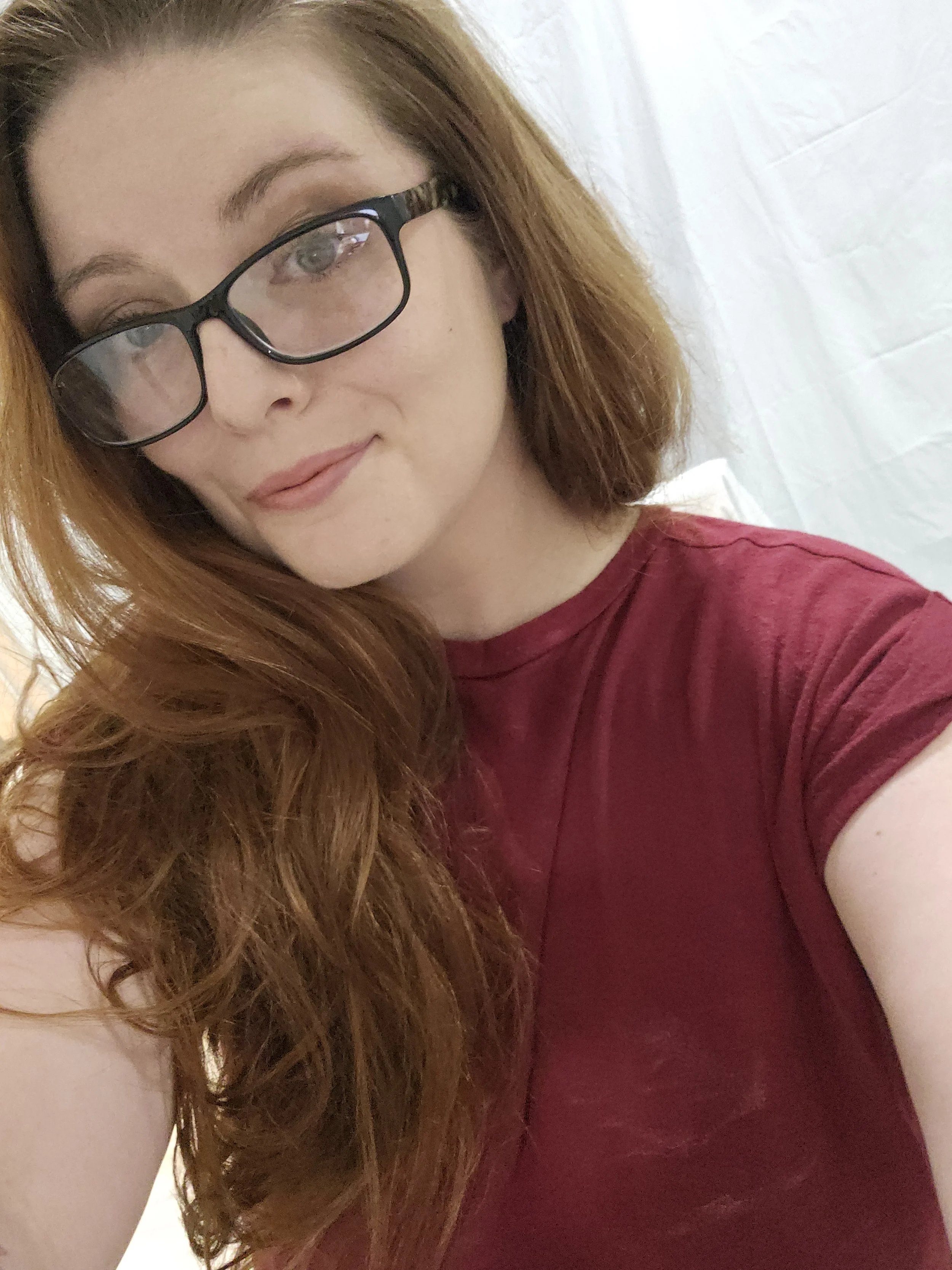 A woman with long red hair wearing glasses and a red shirt, smiling softly, against a plain background.