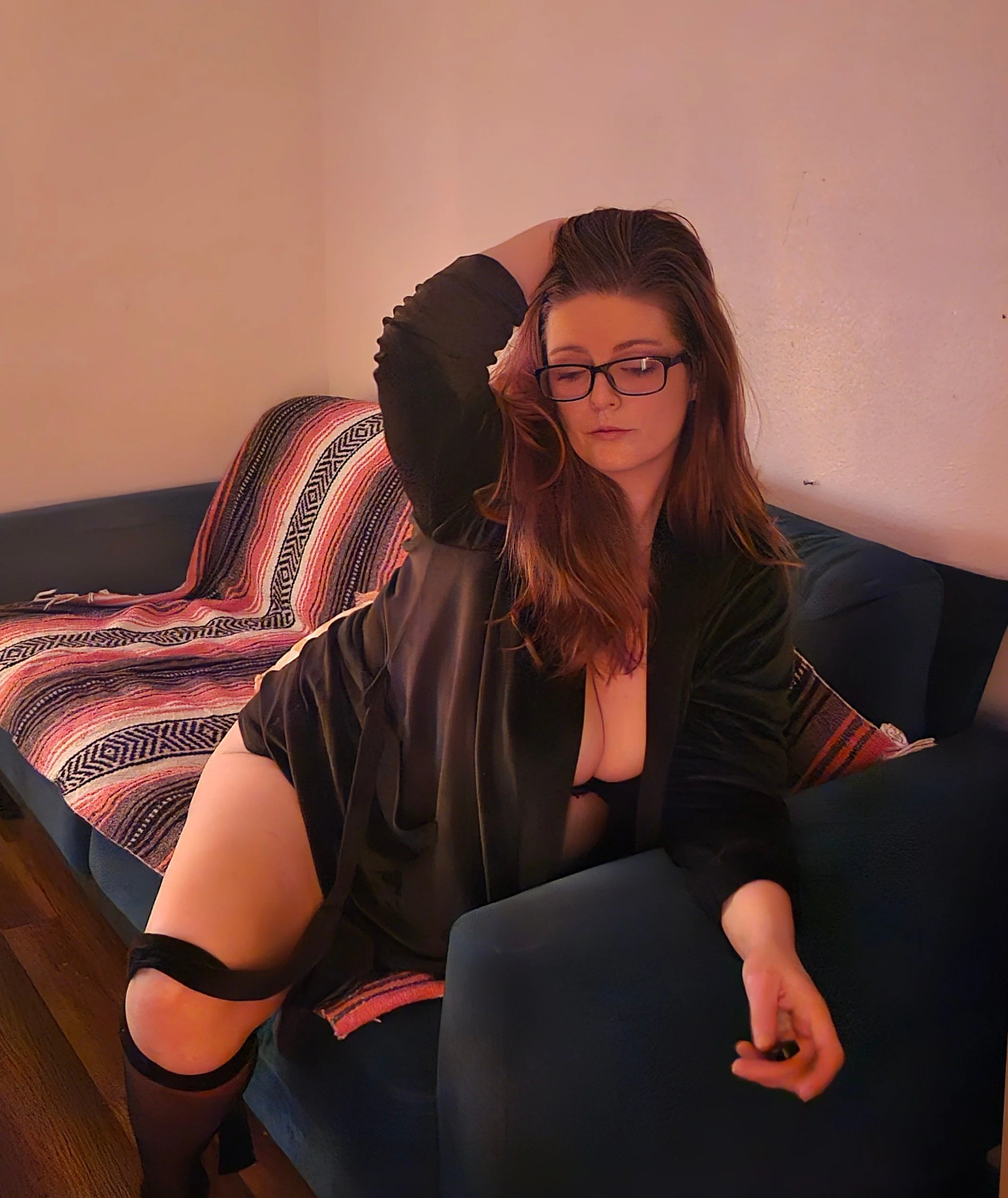 A woman with red hair and glasses sitting on a blue couch, wearing a black robe, with one leg exposed and black stockings, in a dimly lit room.
