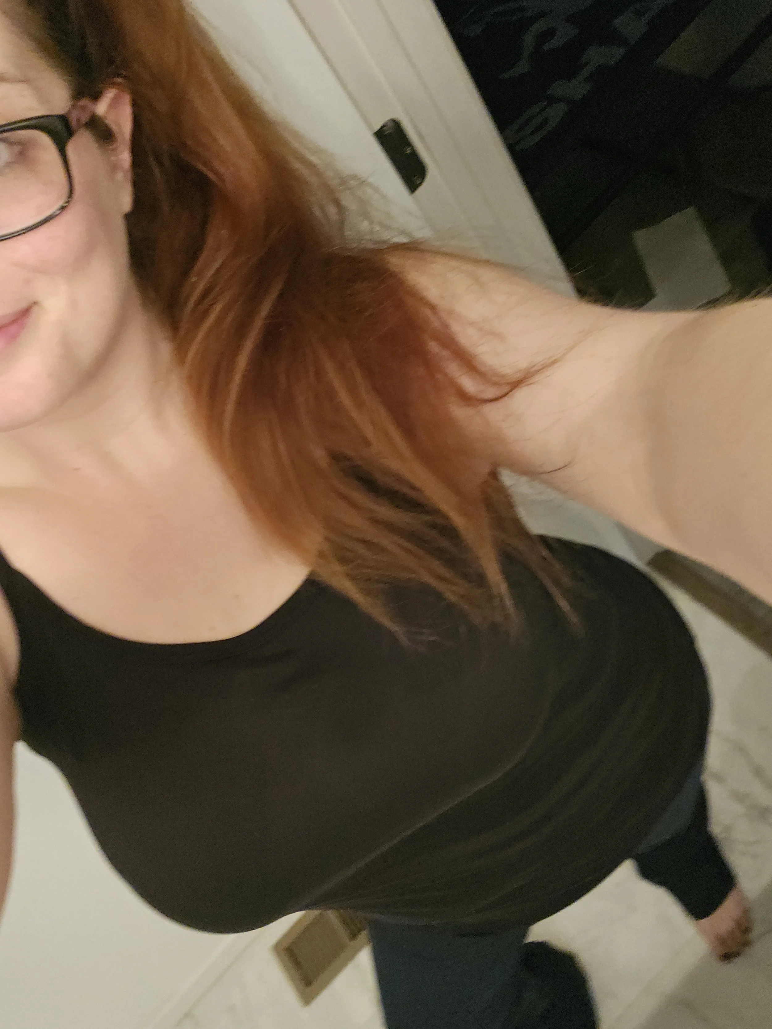 A partial view of a woman with red hair and glasses wearing a black top taking a mirror selfie in a room with a white door and beige walls.