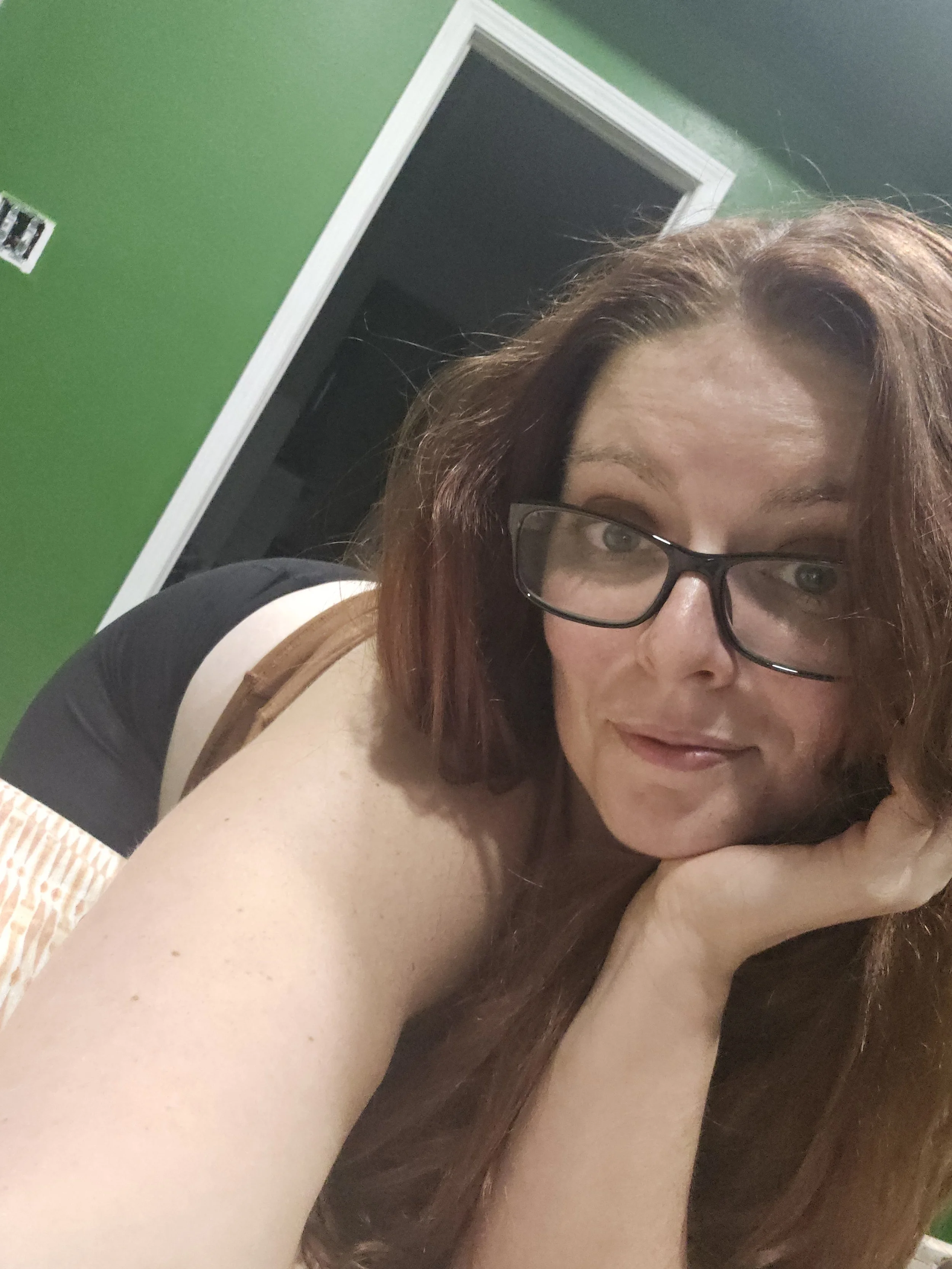 A woman with long red hair and glasses reclining with her head resting on her hand, in front of a green wall with a doorway.