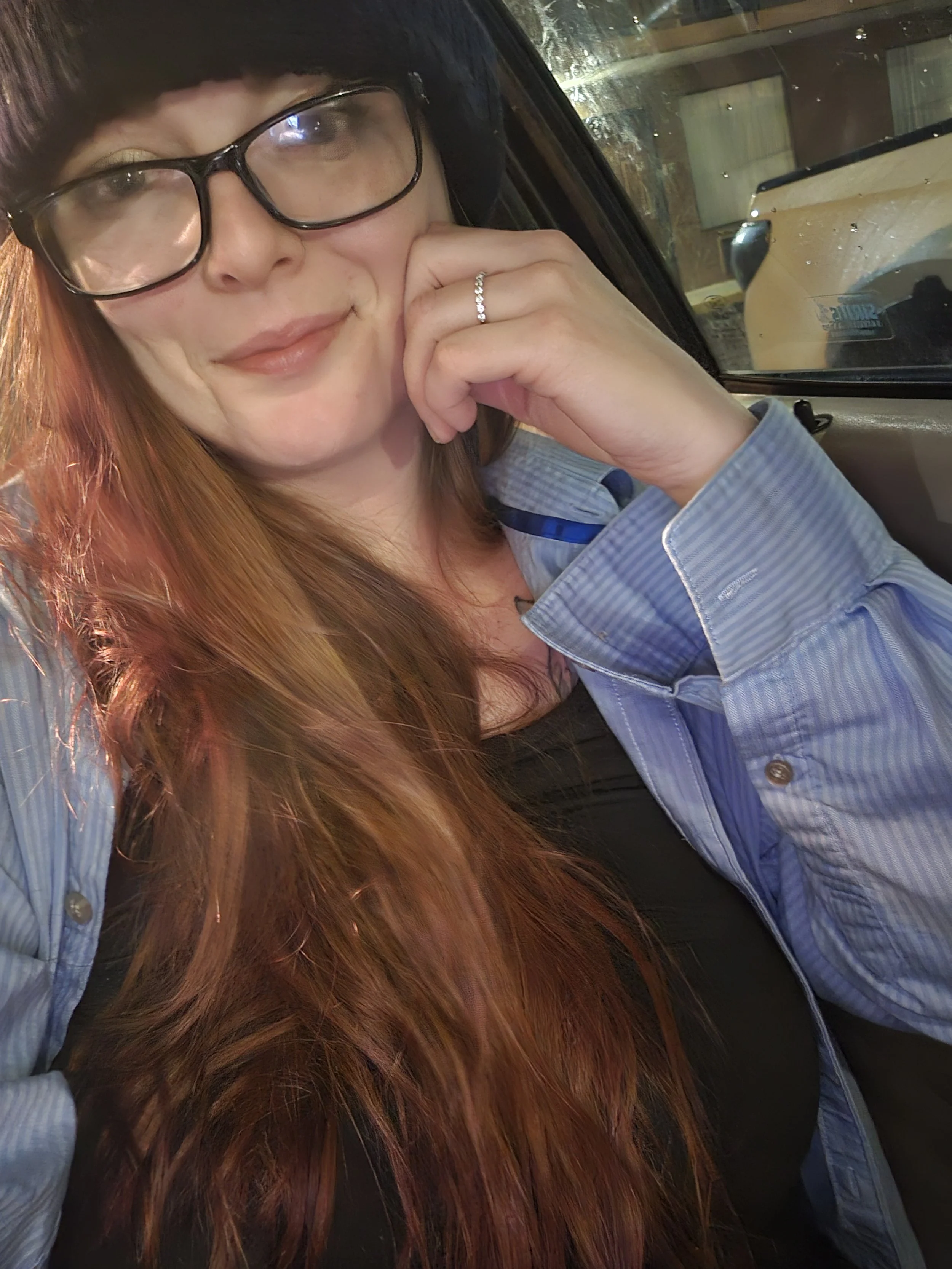A woman with long red hair and glasses sitting inside a vehicle near a window, smiling and resting her chin on her hand, wearing a striped shirt.