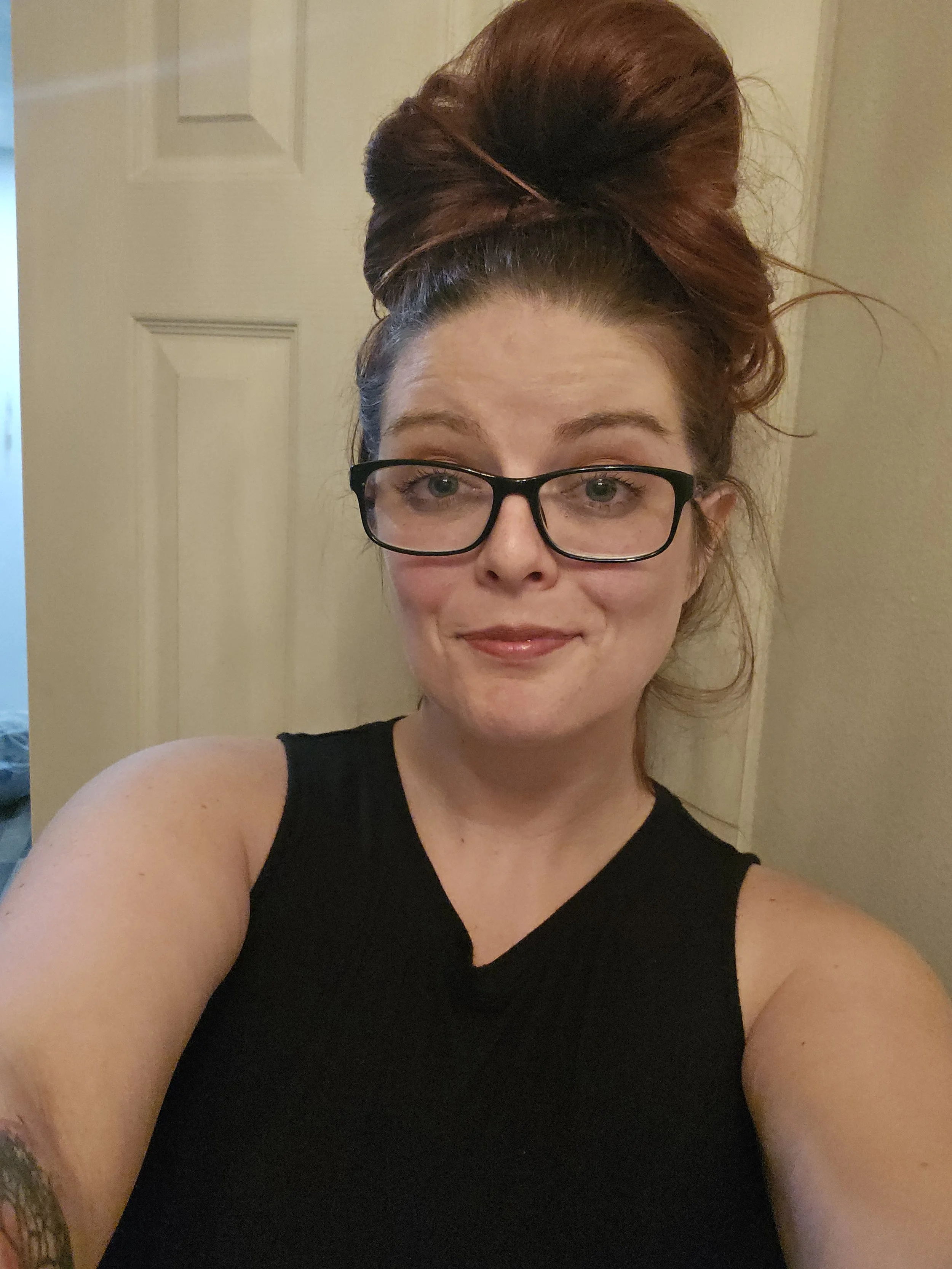 A woman with light skin and glasses, smiling, with her hair up in a large, voluminous bun, wearing a black sleeveless top, standing in front of a beige door.