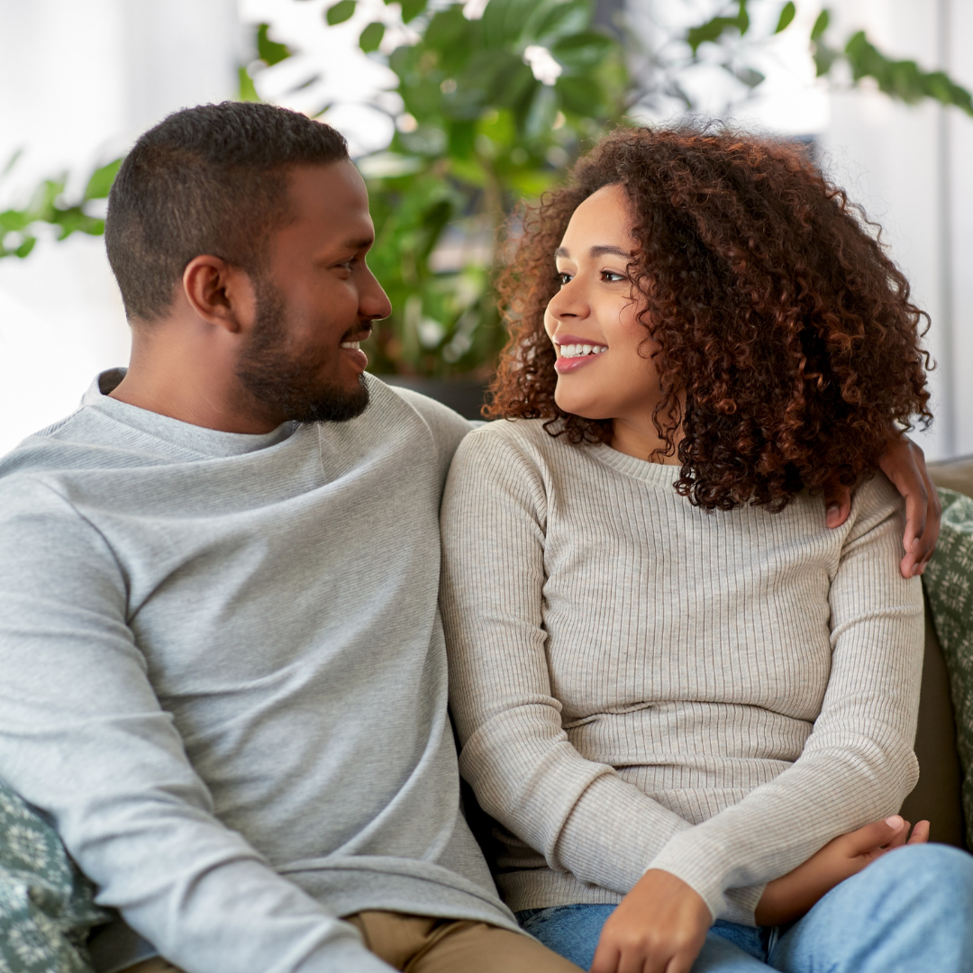 A young couple sitting close together on a couch, smiling and looking at each other affectionately, in a bright living room with green plants in the background.