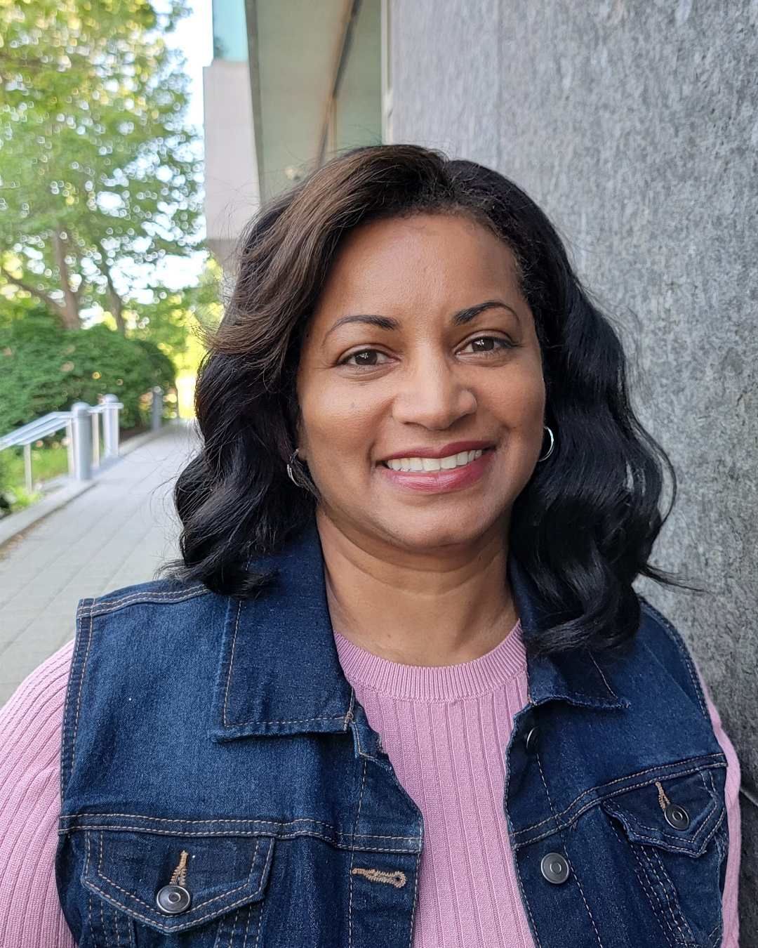 Smiling woman with dark wavy hair wearing a pink sweater and a denim vest outdoors near a gray wall and lush green trees.